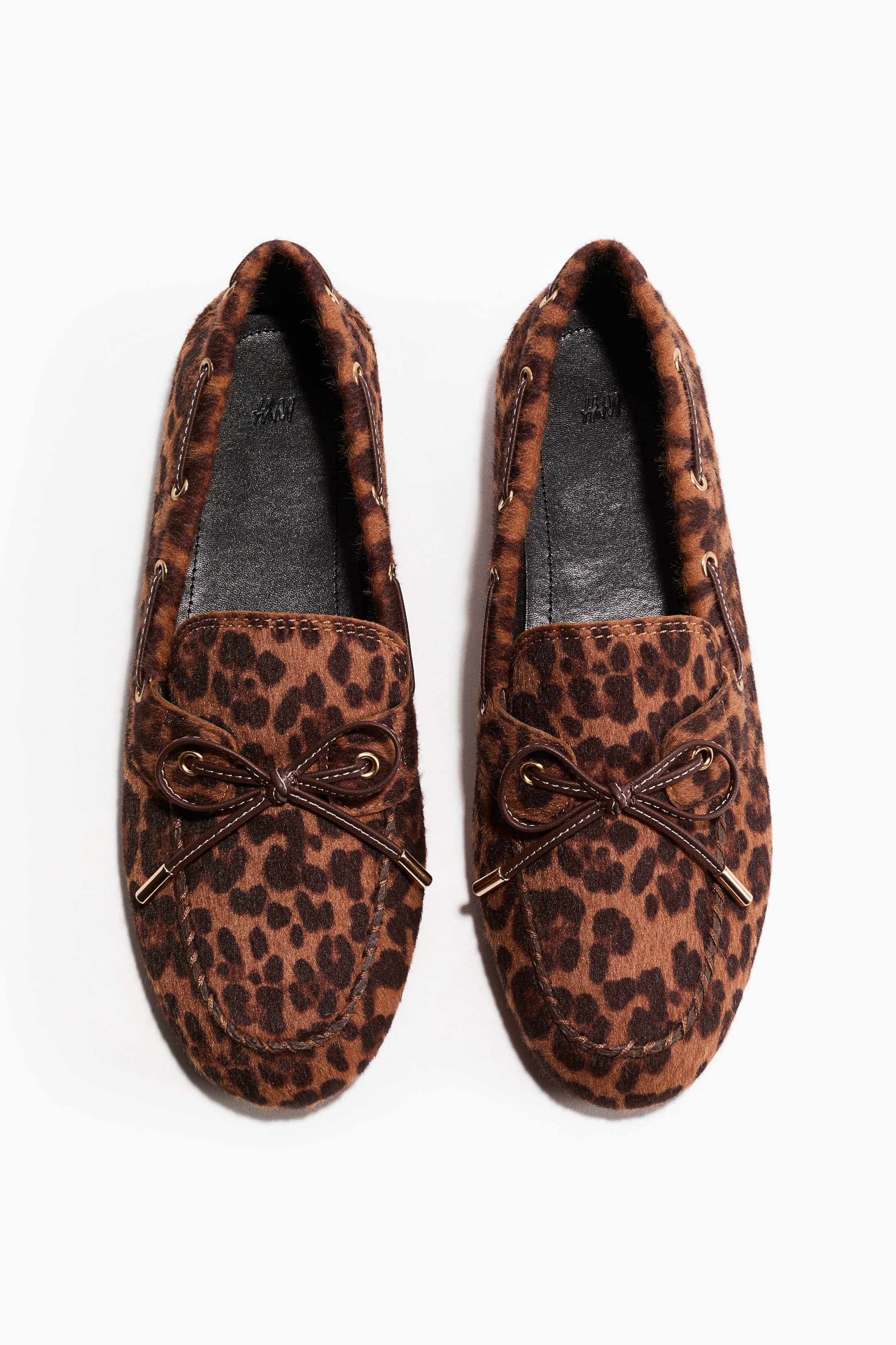 Größeres Bild anzeigen: Leopardenmuster-Slipper stehen frontal und zeigen ein strukturiertes braun-schwarzes Fleckenmuster. Dunkelbraune Schnürsenkel laufen durch goldfarbene Ösen um den Kragen, binden sich zu einer dekorativen Schleife auf dem Rist und enden mit metallischen Spitzen.