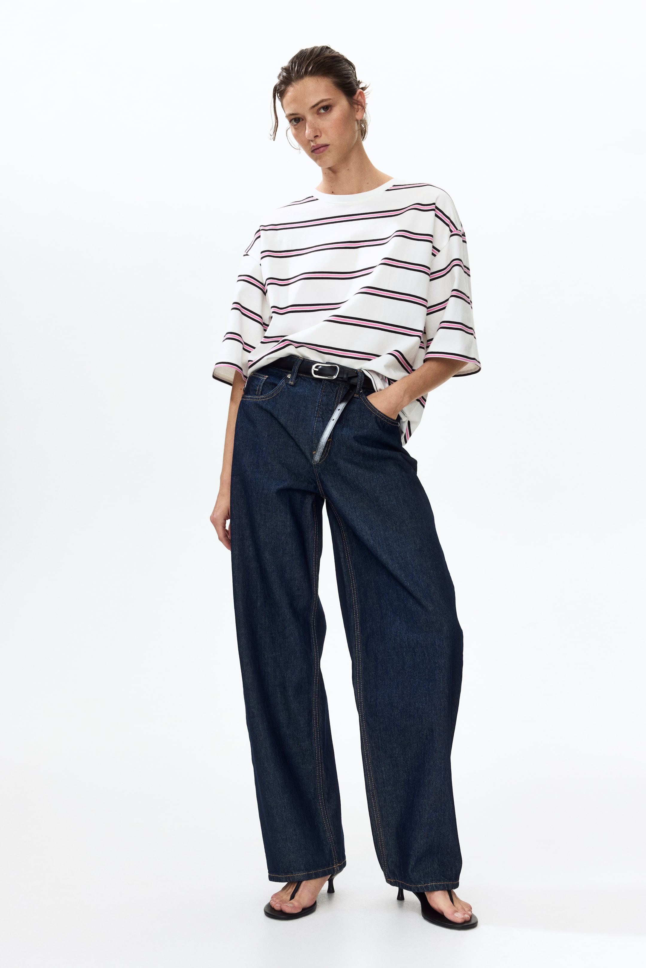 查看大圖: 一件超大號白色T恤，設計有細黑色和粉紅色橫條紋，搭配深色水洗寬腿牛仔褲。牛仔褲由一條帶銀色扣環的黑色皮帶收緊，褲子的全長覆蓋在黑色夾腳涼鞋上。