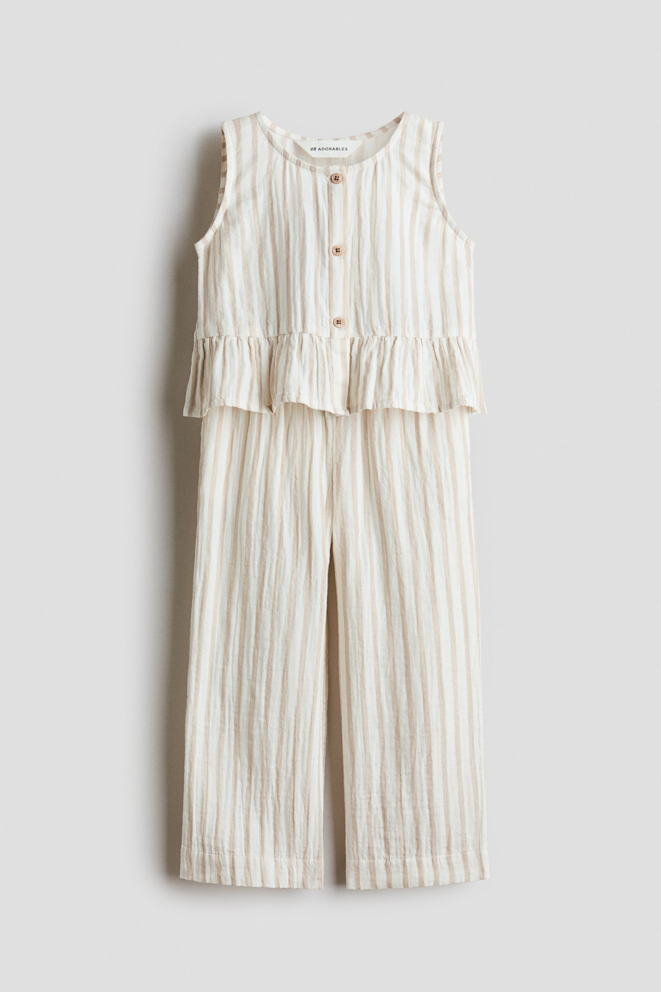 View larger image: A sleeveless jumpsuit featuring a light beige and white vertical stripe pattern on crinkled fabric, shown from the front. It is designed with a round neckline, a front button placket with three brown buttons, a ruffled peplum waist, and wide-leg pants.
