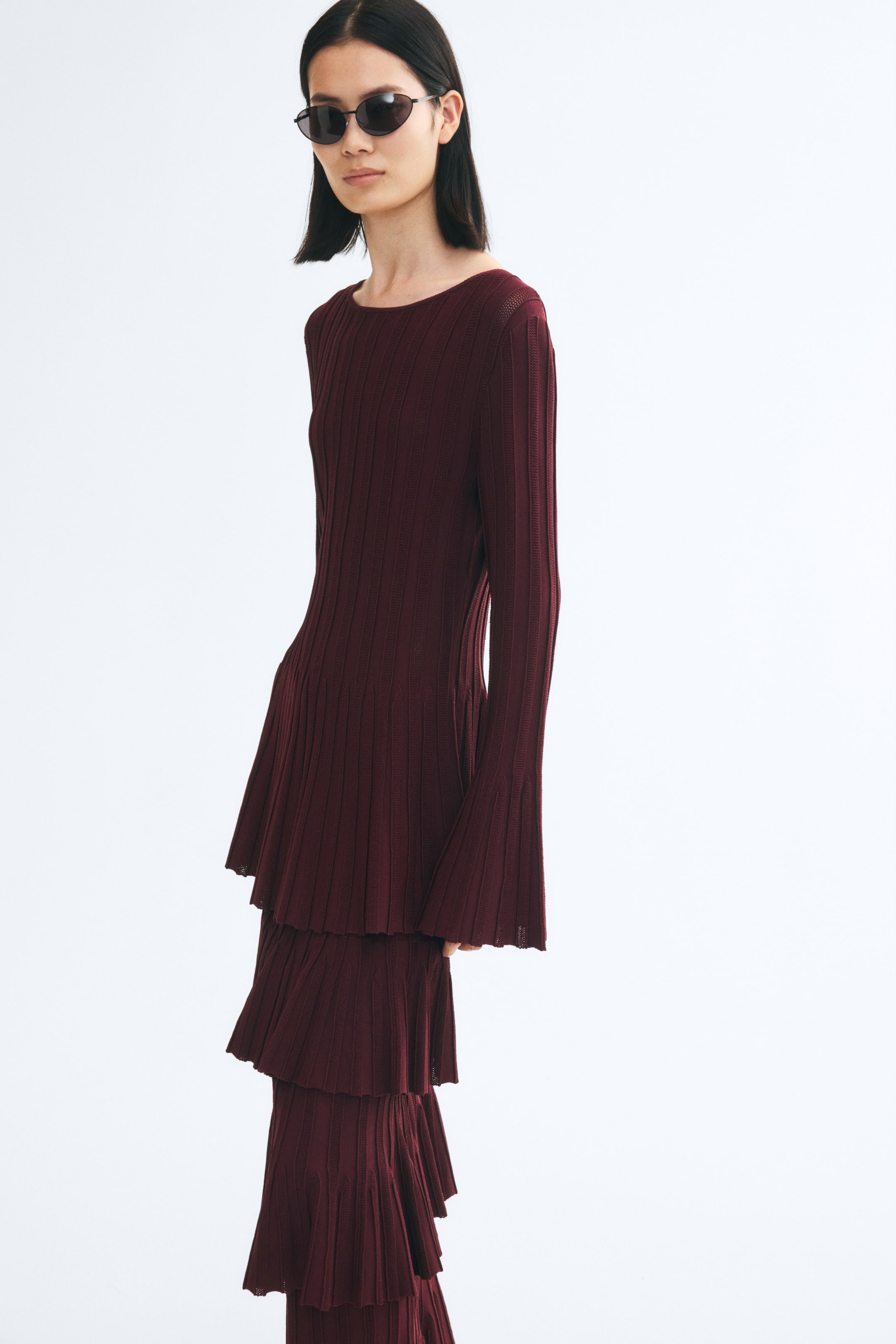 Grotere afbeelding bekijken: Het model draagt een bordeauxrode top in ribgebreide stof, met een aansluitend silhouet, een ronde hals, lange mouwen met uitlopende manchetten en een licht uitlopende peplumzoom. De top wordt gestyled met een bijpassende gelaagde ribgebreide rok en donkere zonnebril.