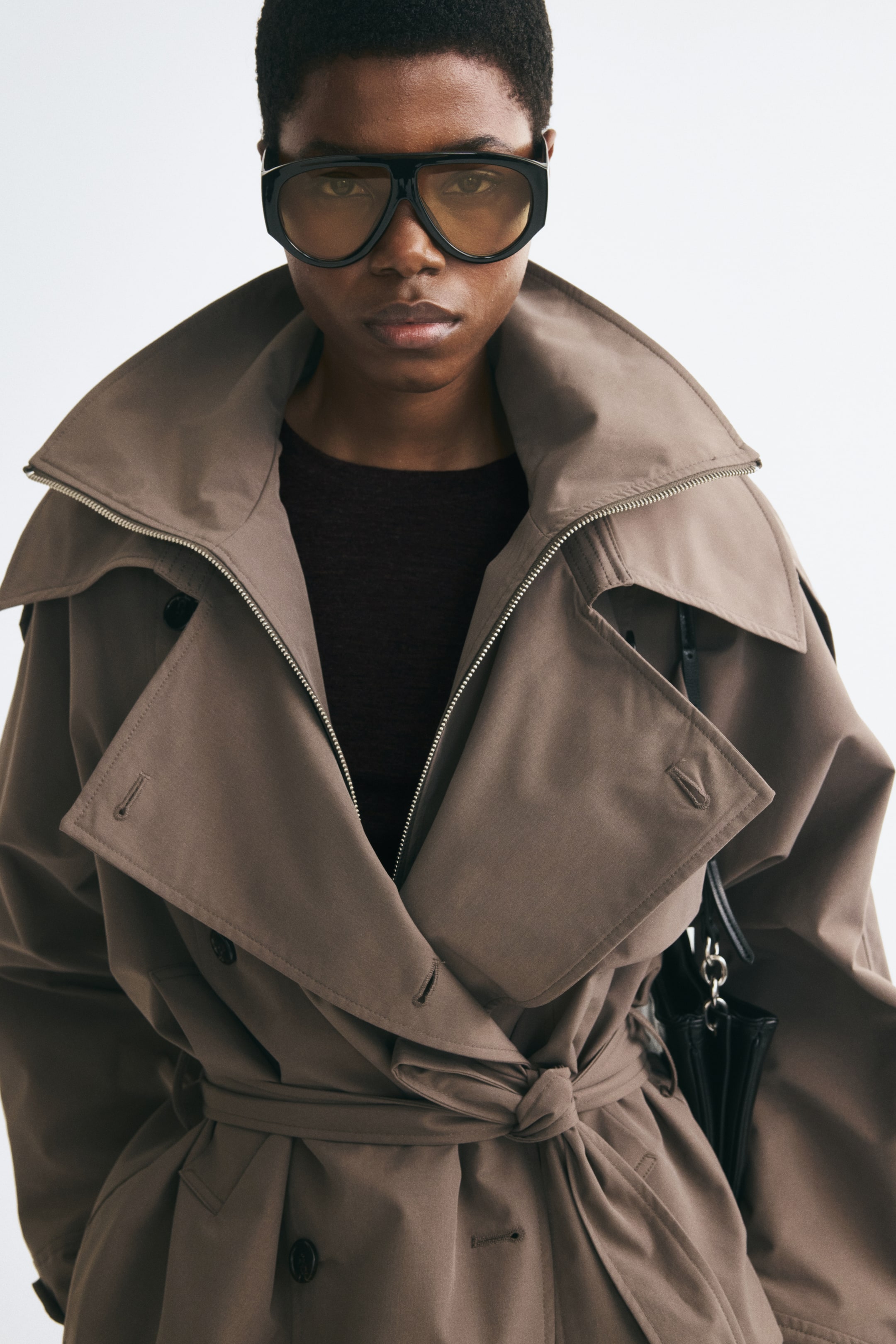 Visualizza immagine più grande: Una maglia aderente marrone scuro in lana a fine maglia viene indossata dalla modella come strato base sotto un trench strutturato color taupe. Il top presenta un girocollo rifinito a costine, visibile nella zona del petto dove il cappotto è portato aperto.