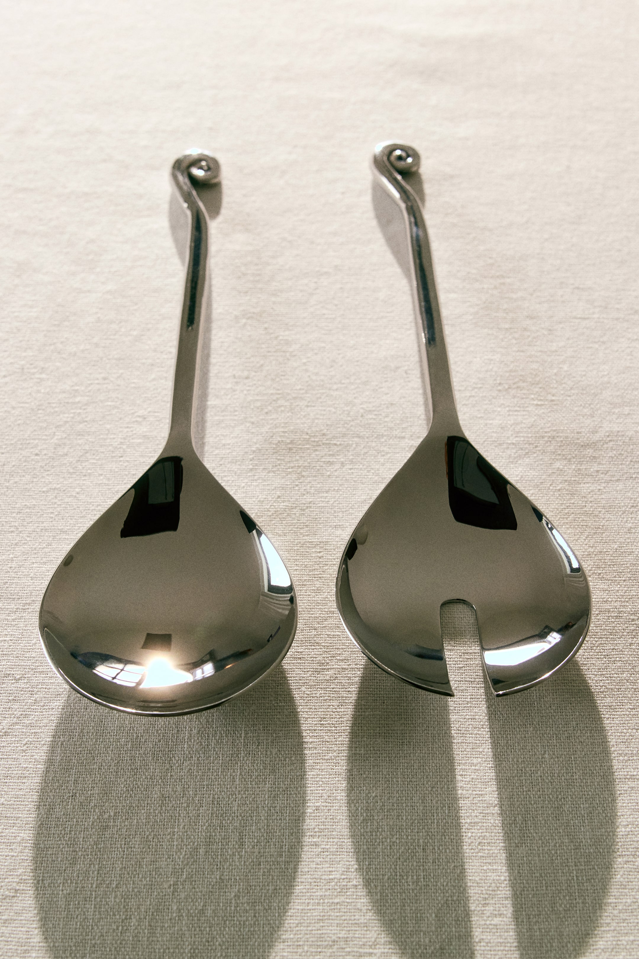 Ver imagen más grande: Dos cubiertos de ensalada de metal pulido están colocados uno al lado del otro, ligeramente inclinados hacia arriba, cada uno con un mango largo que culmina en un extremo decorativo en espiral. Uno de los cubiertos presenta un cuenco sólido en forma de lágrima, mientras que el otro muestra un cuenco a juego en forma de lágrima con una ranura central.