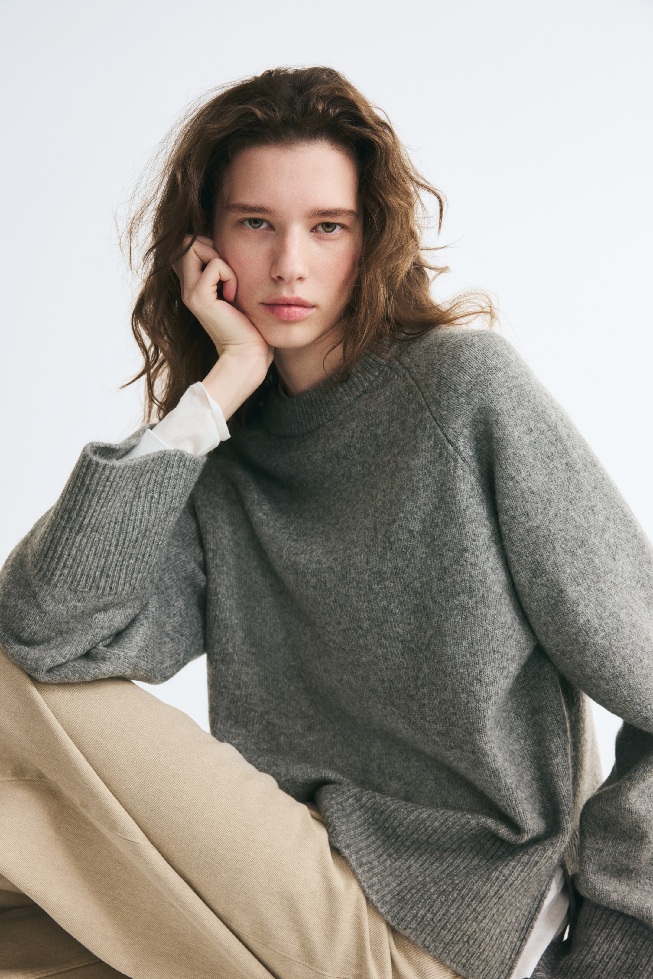 Voir une image plus grande: Un chandail gris en tricot doux, doté d’un col rond bordé de côtes, de manches raglan longues et de larges poignets côtelés, est porté par le mannequin. Le chandail est superposé à une chemise blanche à manches longues et agencé avec un pantalon beige.