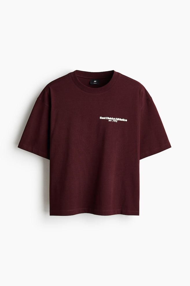 前面から見ると、オーバーサイズでゆったりとしたシルエットを持つマルーンのボクシースタイルTシャツです。丸いリブトリムのネックライン、ドロップショルダー、そして左胸に白い「East District Athletics est. 1999」の文字がプリントされています。 - 1