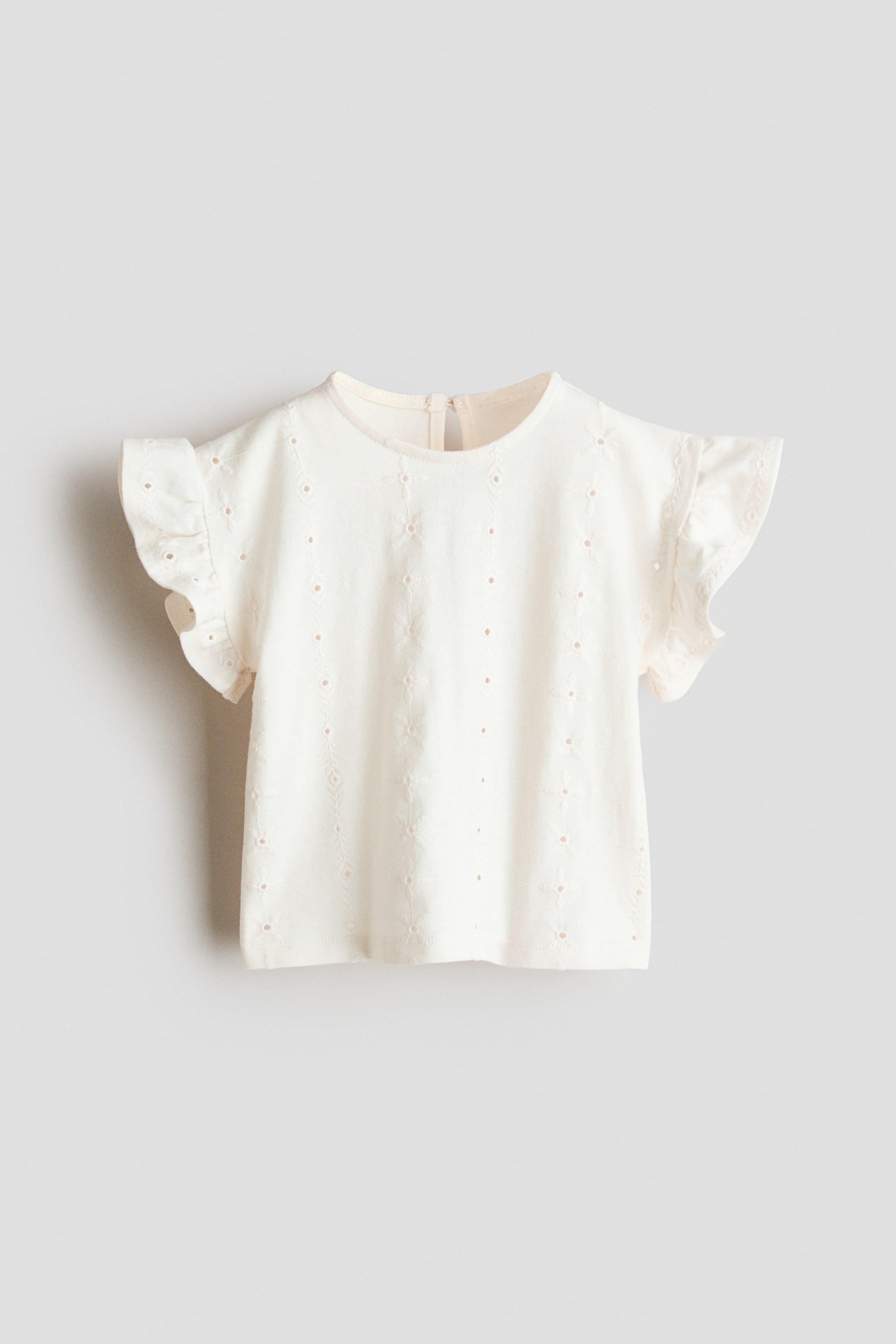Agrandir l'image: Le haut crème présente un col rond et des manches courtes à volants. Il est orné d'une broderie anglaise avec de petits motifs floraux et étoilés sur le devant, et une petite ouverture en goutte d'eau avec un fermoir est visible à l'arrière du col.