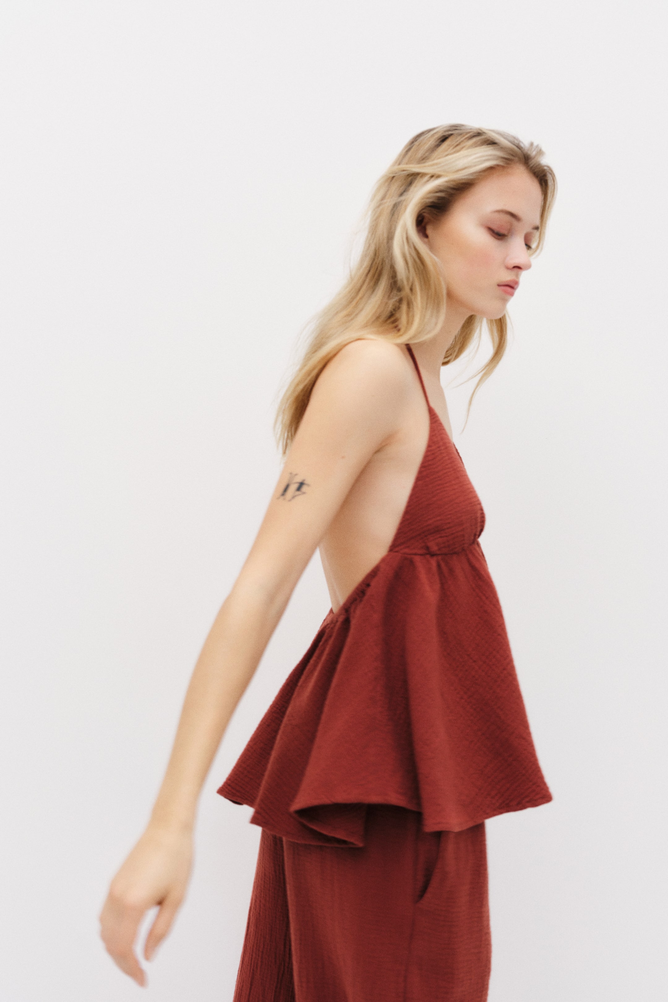 View larger image: The rust-coloured top is showcased from a side-back angle, highlighting its textured fabric that shapes a fitted bodice and a flared peplum hem. The design of the top reveals a low-cut back, complemented by matching textured trousers worn below.