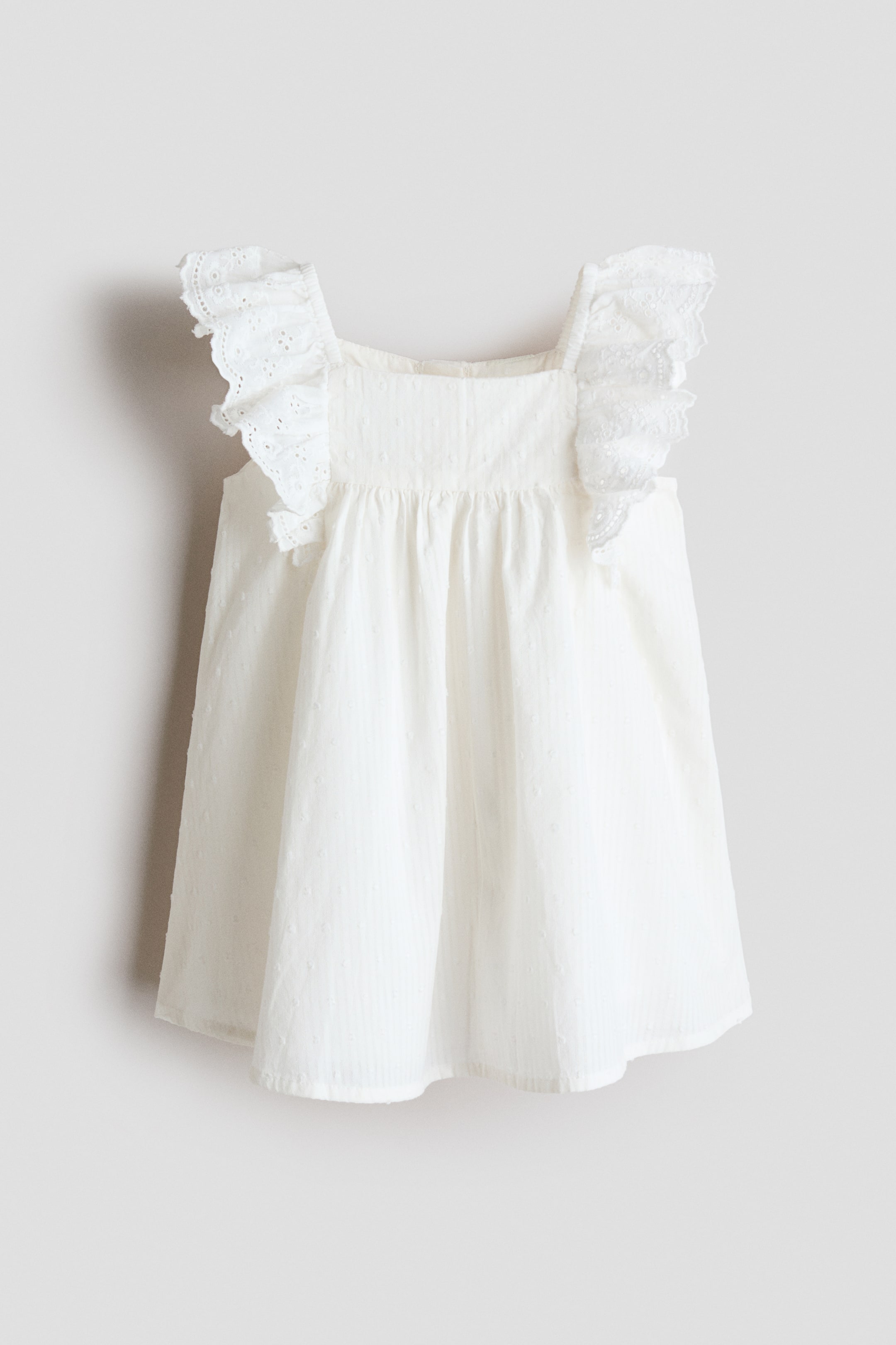 Vis større bilde: En hvit baby doll-kjole presenteres foran, med en firkantet utringning og rynkede detaljer under livet. De brede stroppene er pyntet med broderte blonderysjer, og stoffet har en subtil tekstur med små hevede prikker.