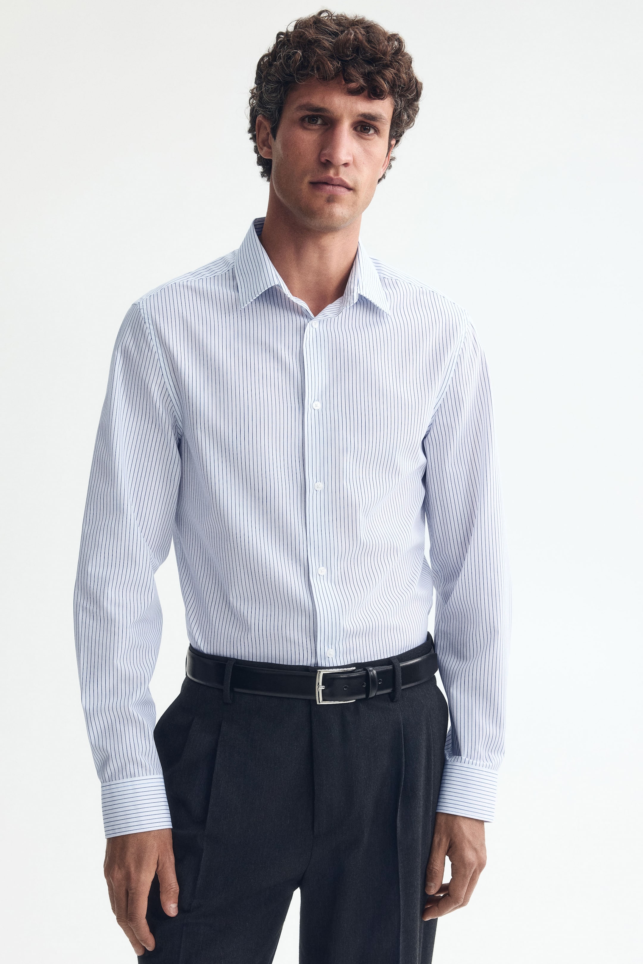 Ver imagem maior: Uma camisa branca de corte justo com finas riscas verticais azuis está a ser usada pelo modelo, apresentando uma gola clássica, frente com botões e mangas compridas com punhos abotoados. A camisa está enfiada em calças escuras, criando uma silhueta ajustada.