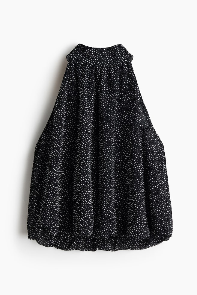 Un top sans manches à col halter en tissu tissé noir à pois blancs est suspendu, vu de face, présentant un col montant froncé et une silhouette volumineuse qui retombe en un ourlet boule bouffant. - 1
