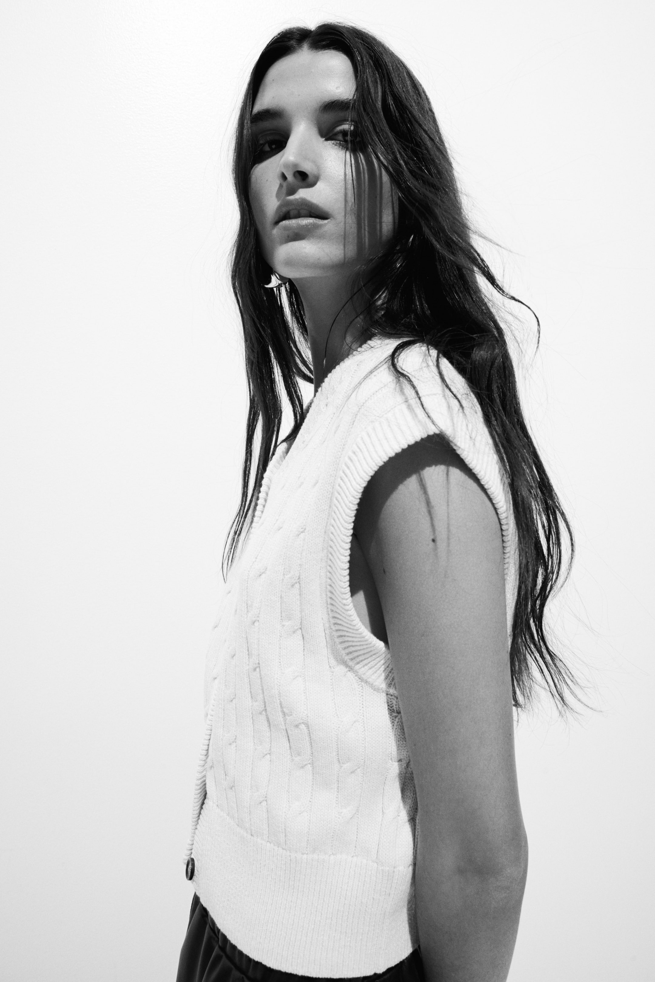 View larger image: A white cable-knit jumper vest is being worn, angled slightly to the right, featuring a V-neckline, a cable-knit pattern, and a ribbed hem. A dark button closure is visible on the left side near the hem.