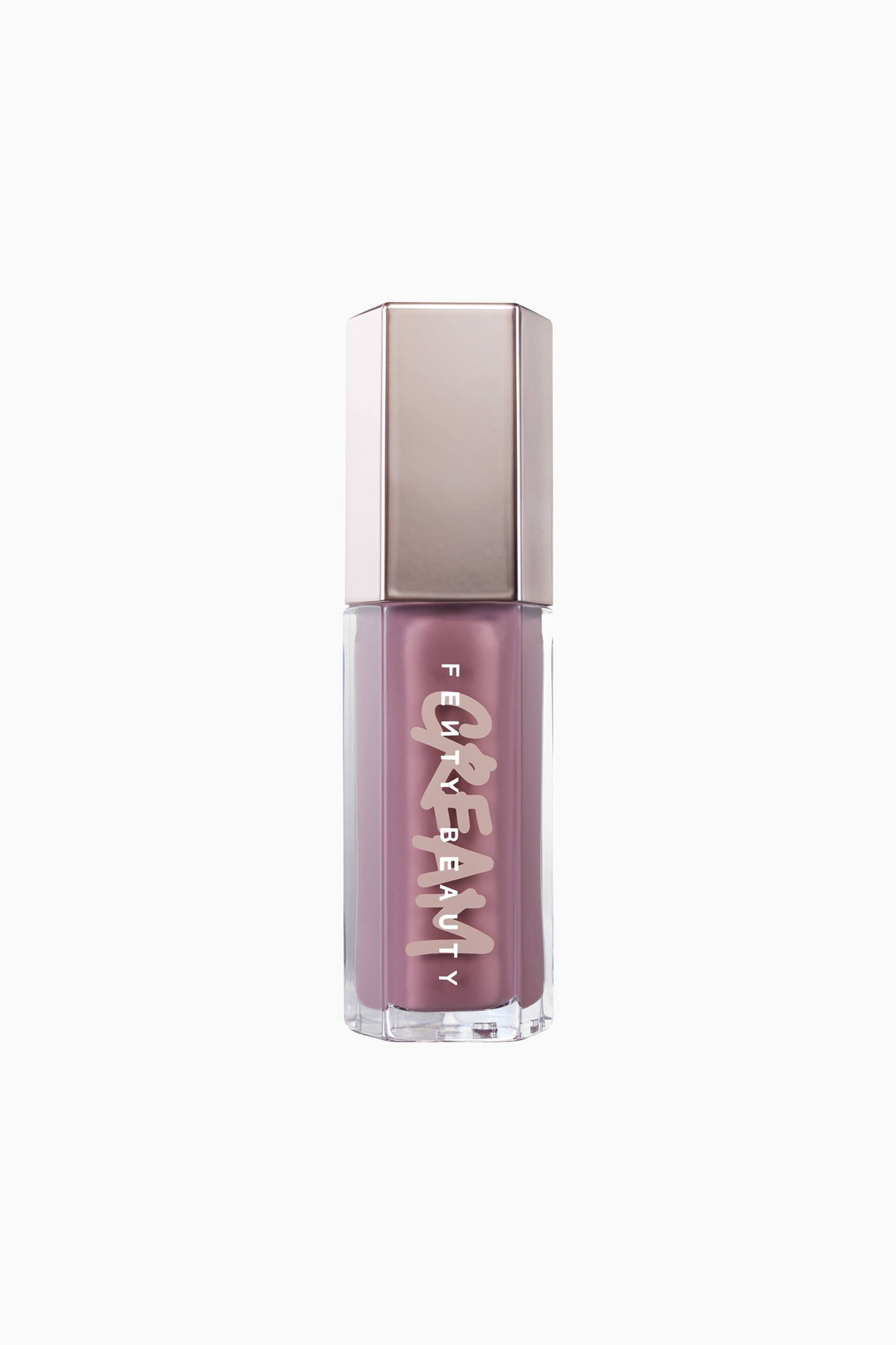 Näytä suurempi kuva: Kuusikulmainen Fenty Beauty Gloss Bomb Cream -huulikiiltoputkilo seisoo pystyssä, ja sen kirkkaan säiliön läpi näkyy pölyinen mauve-vaaleanpunainen nestemäinen koostumus. Pullon päällä on heijastava, metallinhohtoinen ruusukultainen kuusikulmainen korkki, ja siinä on brändin nimi "FENTY BEAUTY" painettuna pystysuoraan tyylitellyn, vaaleanpunaisen "CREAM"-logon yläpuolelle.