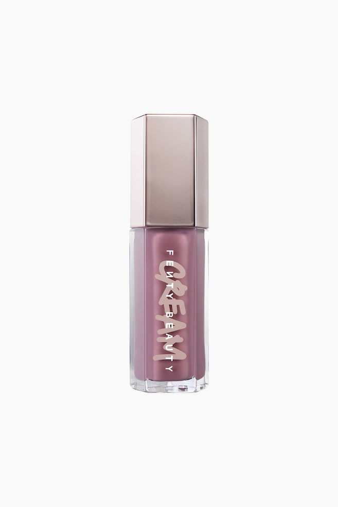 Et sekskantet Fenty Beauty Gloss Bomb Cream lÃ¦begloss-rÃ¸r stÃ¥r oprejst og fremhÃ¦ver en stÃ¸vet mauve-rosa flydende formel gennem den klare beholder. Flasken er toppet med en reflekterende, metallisk rosaguld sekskantet hÃ¦tte og har brandnavnet "FENTY BEAUTY" trykt lodret over et stiliseret, lyserÃ¸dt "CREAM"-logo. - 1