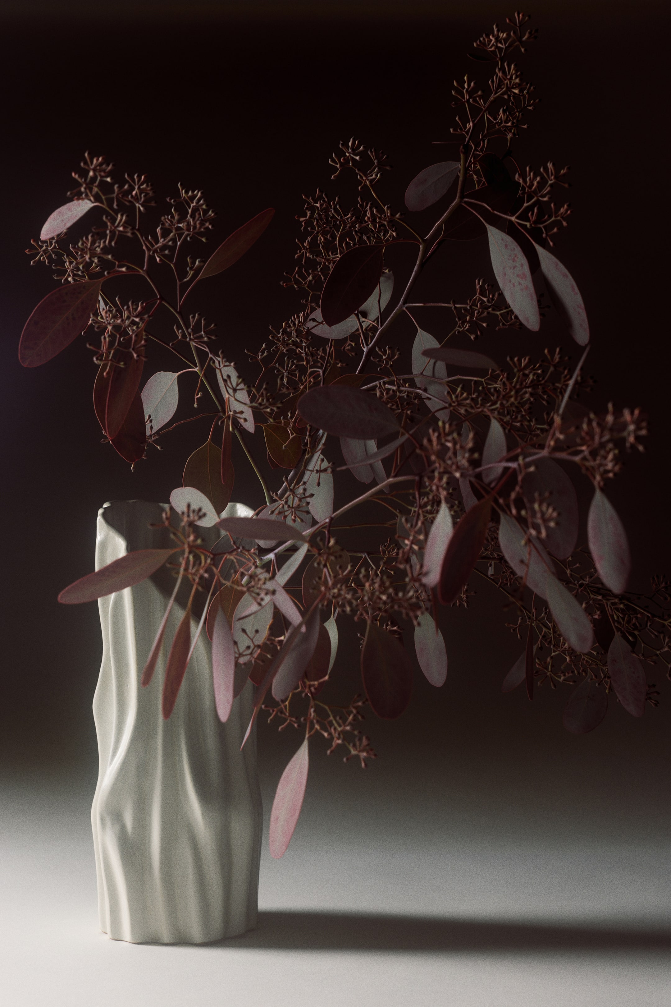Agrandir l'image: Un vase sculptural en céramique blanc cassé se tient debout, présentant une surface texturée et ondulée avec des stries verticales et un bord asymétrique. Le vase contient des branches d’eucalyptus séchées, brun rougeâtre, avec de petites capsules de graines et des feuilles ovales retombant sur le côté.