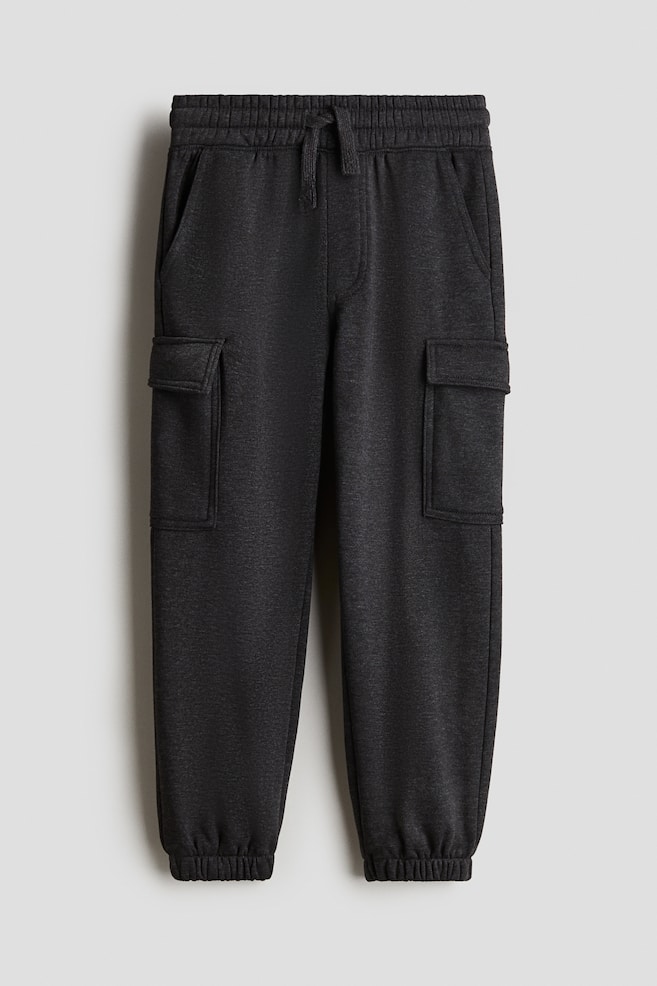 A pair of dark grey cargo joggers is being shown from the front. They are featuring an elasticated waist with a decorative drawstring, and diagonal side pockets are visible. Large flap pockets are positioned on the legs, and covered elastication is gathering the hems. - 1