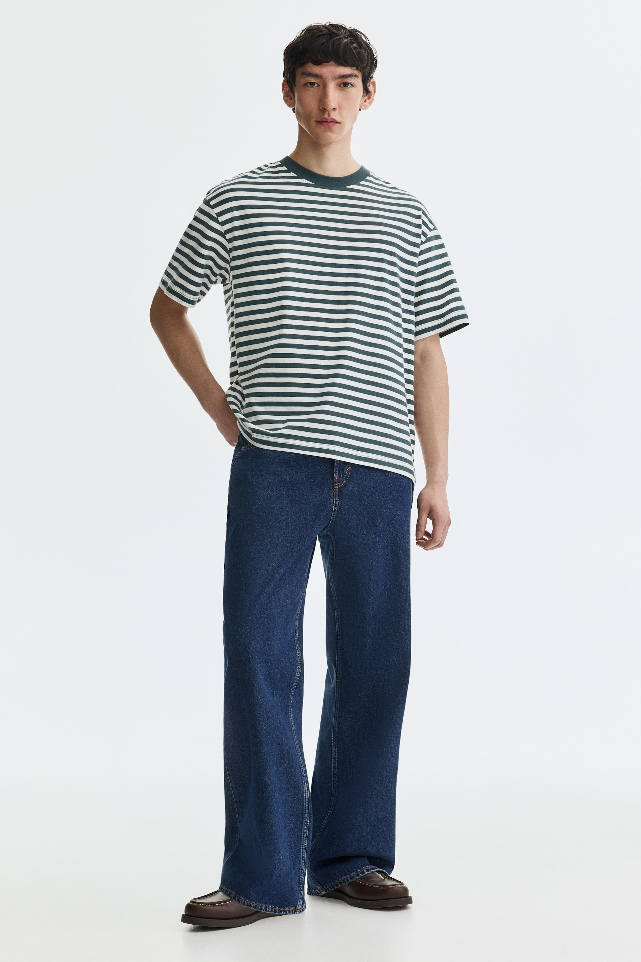 Grotere afbeelding bekijken: Een donkergroen en wit gestreept t-shirt met een geribbelde ronde hals en korte mouwen. Het wordt gecombineerd met donkerblauwe wijde jeans, die zichtbare oranje stiksels en een ontspannen pasvorm tonen, gepresenteerd vanuit een lichte voor-linker hoek.