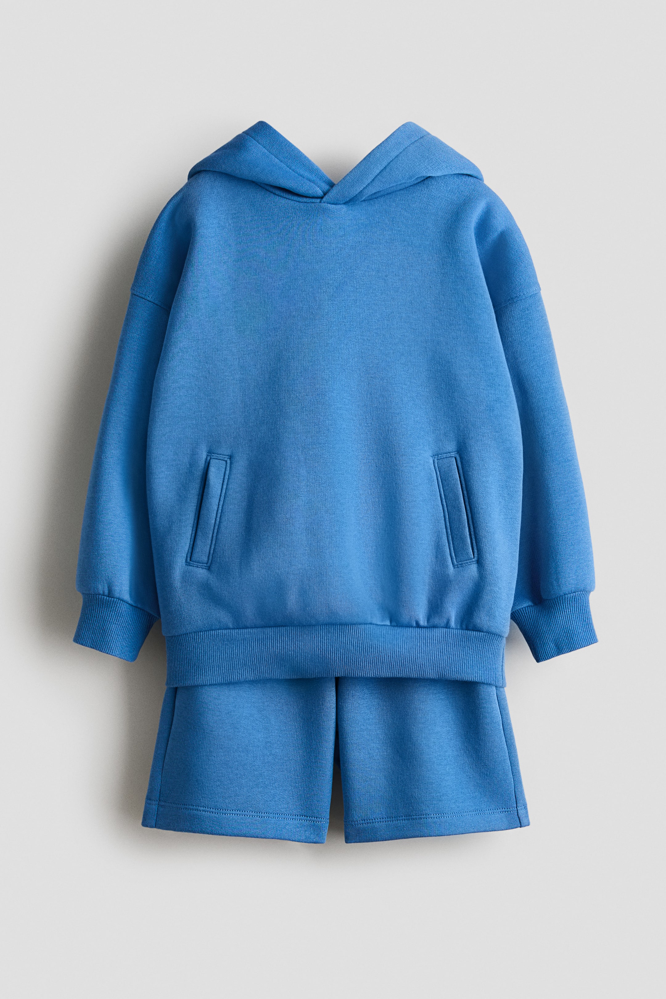 Ver imagem maior: Um conjunto de hoodie azul e calções é apresentado de frente. O hoodie apresenta um capuz, mangas compridas com punhos canelados, uma bainha canelada e dois bolsos de vivo retangulares. Os calções azuis combinantes estão visíveis por baixo do hoodie, com uma bainha lisa.