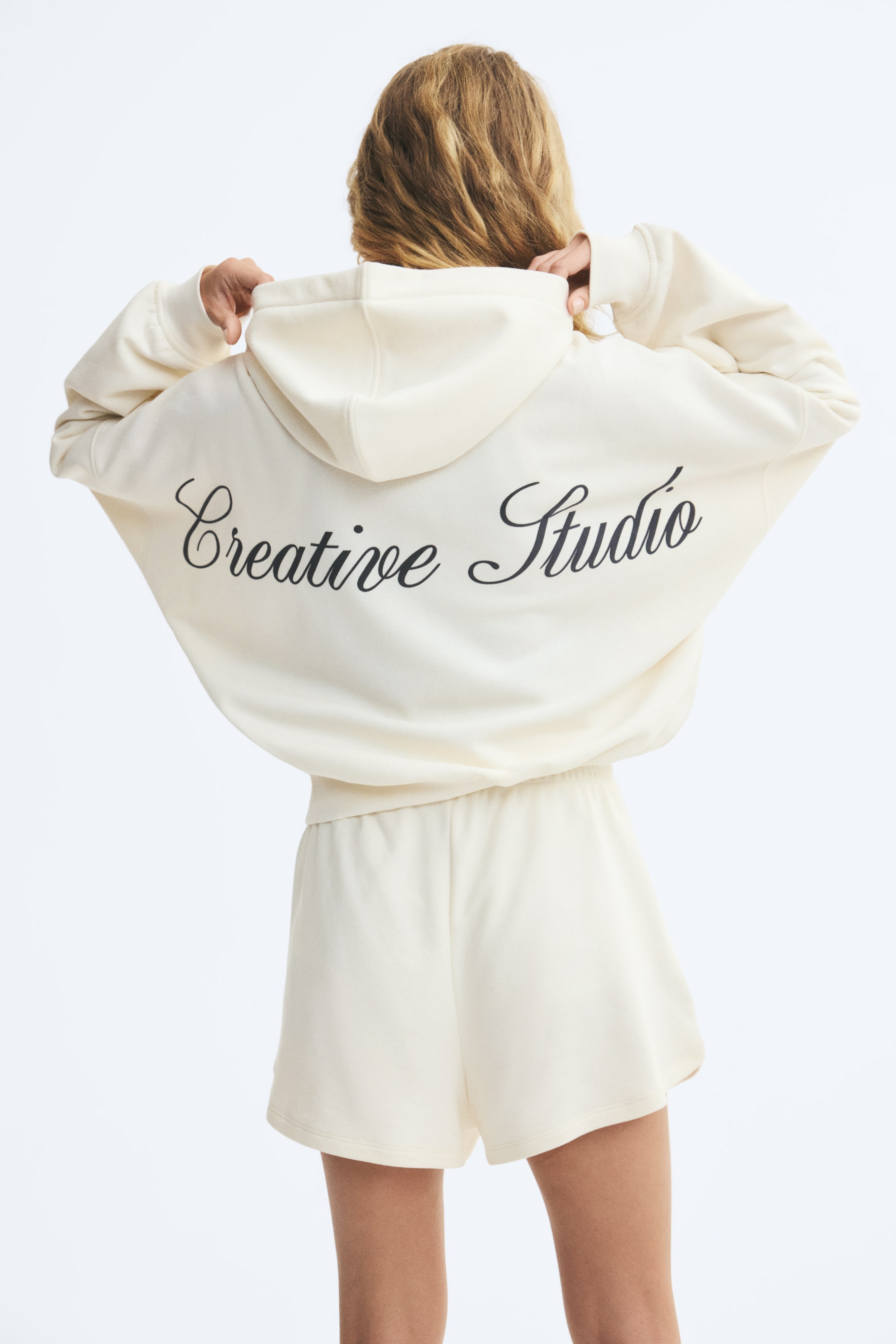View larger image: An off-white, relaxed-fit hoodie featuring black script text reading "Creative Studio" across the back is being worn by the model, who is holding the sides of the hood. The hoodie features dropped shoulders and a ribbed hem, and is paired with matching off-white shorts.