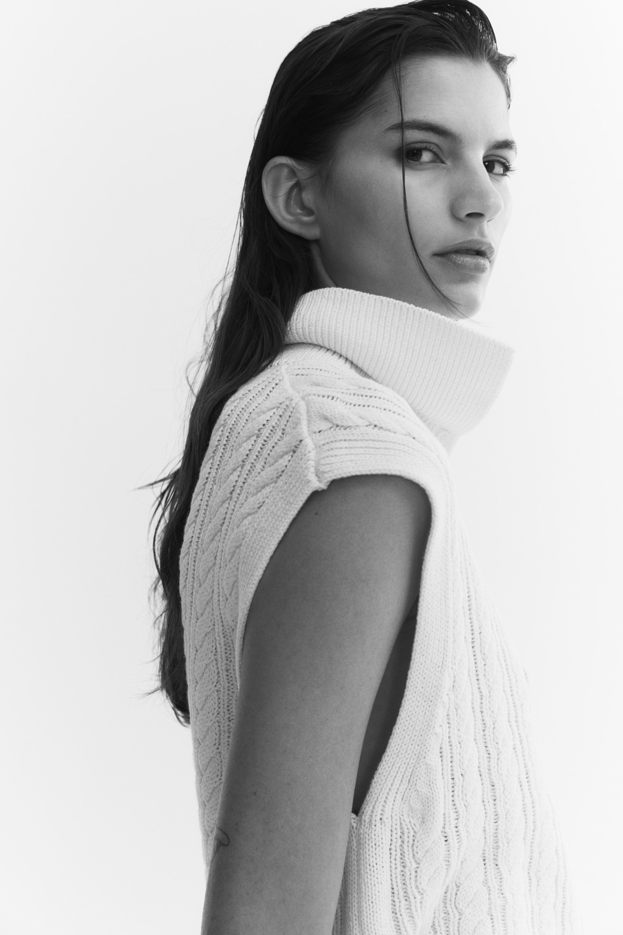 Ampliar la imagen: Una chaleco de punto trenzado con cuello alto acanalado tipo tortuga lo lleva la modelo en una vista de perfil lateral. La prenda sin mangas muestra un patrón trenzado texturizado en el cuerpo y ribetes acanalados en las sisas.