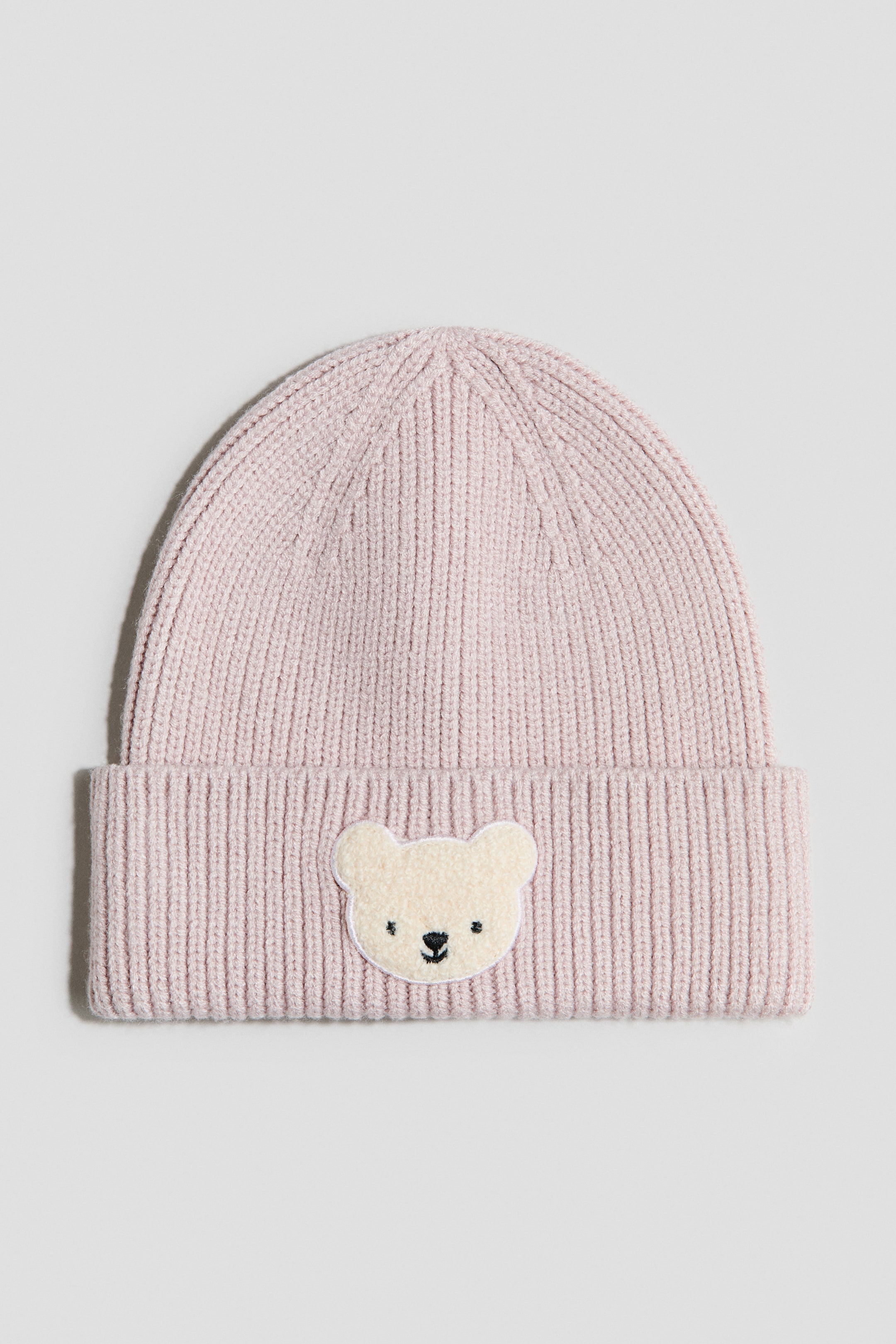 View larger image: A light pink ribbed knit beanie is lying flat, featuring a cream-coloured bear head patch with embroidered black eyes and nose on its folded cuff.