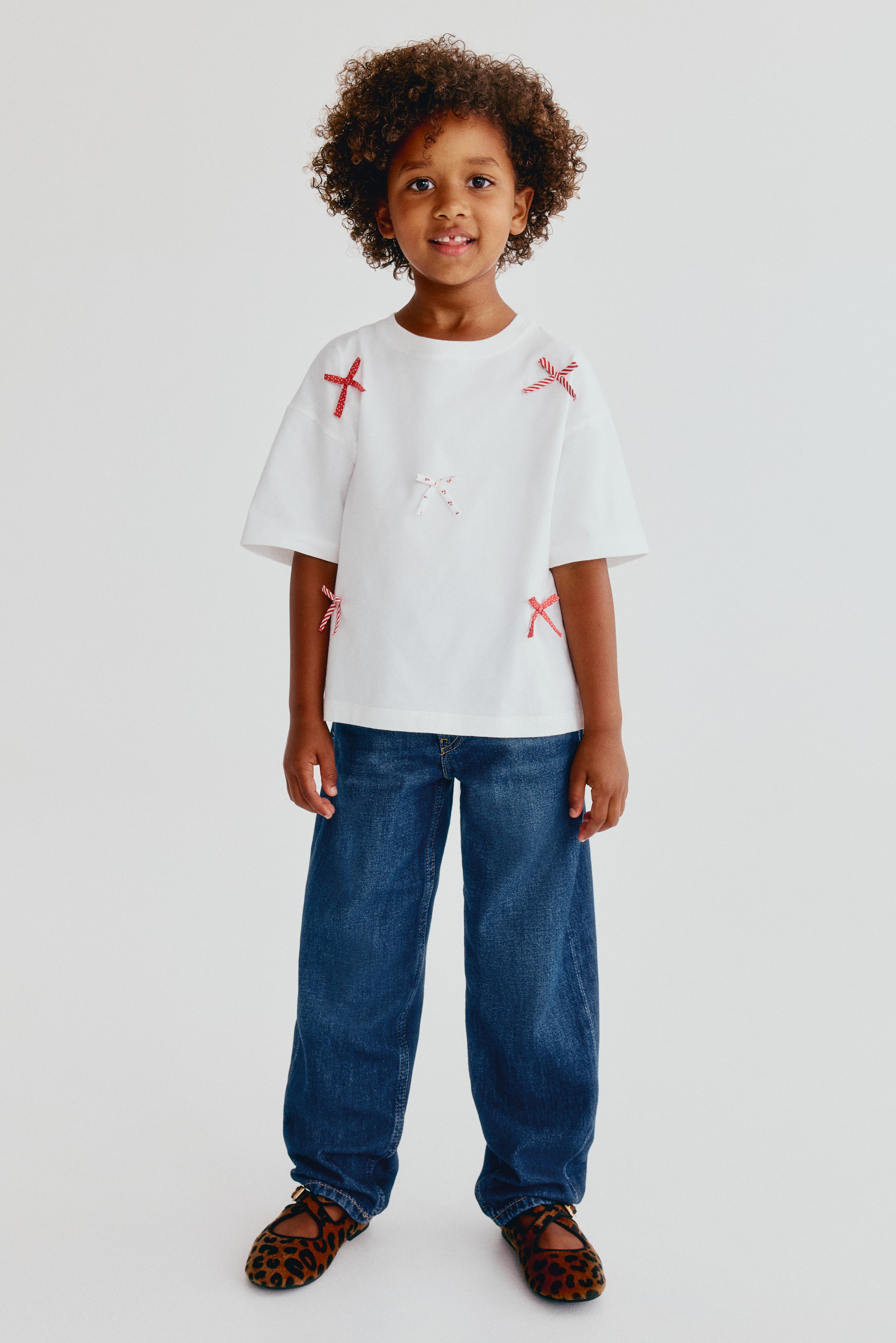 Ver imagem maior: Uma criança está a vestir estes jeans de ganga azul em algodão de corte largo, mostrados de frente, com um corte descontraído nas coxas e pernas ligeiramente afuniladas. O cós dos jeans está coberto por uma t-shirt branca.