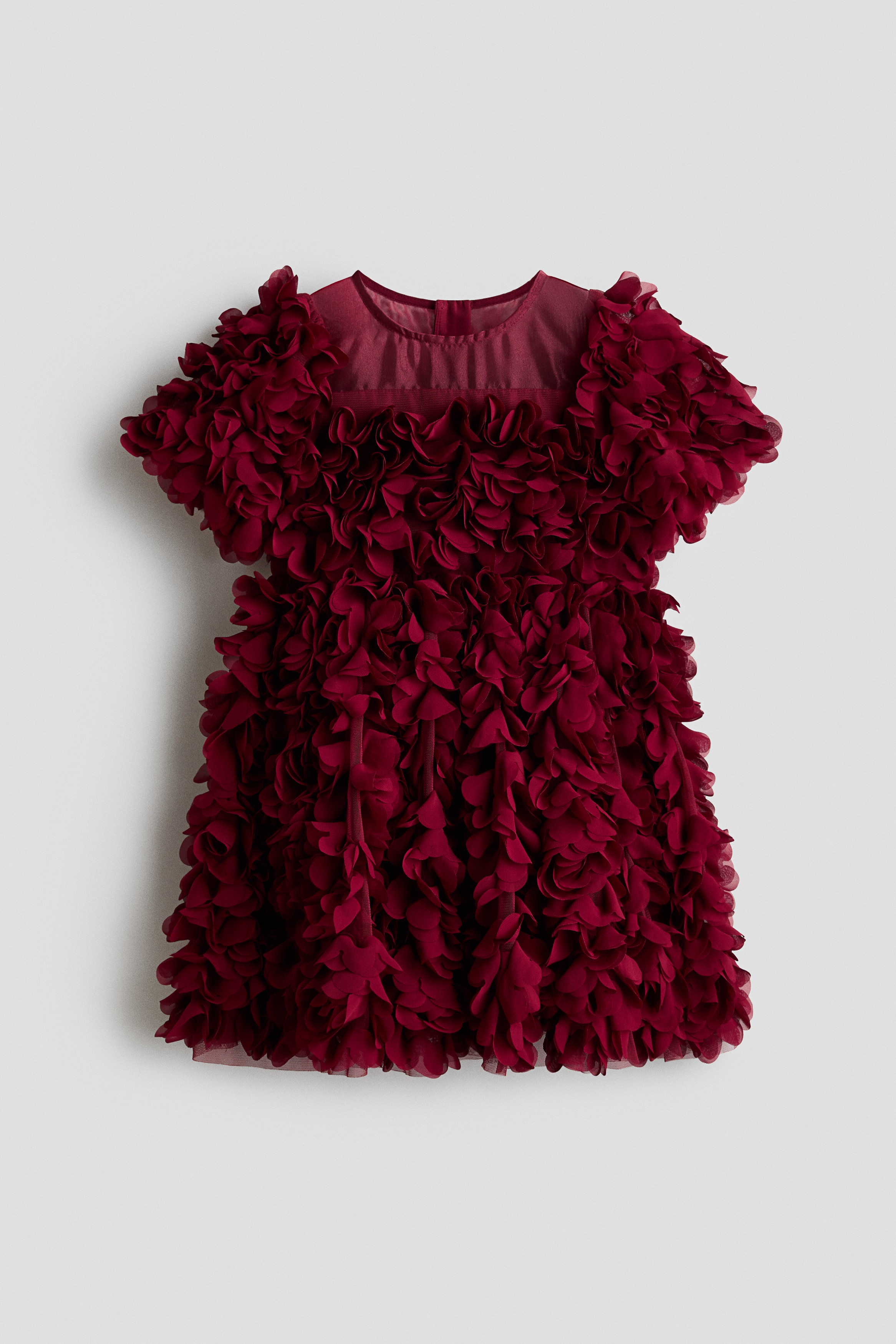 A dark red dress is shown from a side angle, featuring dense, three-dimensional fabric ruffles that cover the entire garment from the short sleeves to the flared skirt. The dress presents a highly textured, voluminous design while being worn.