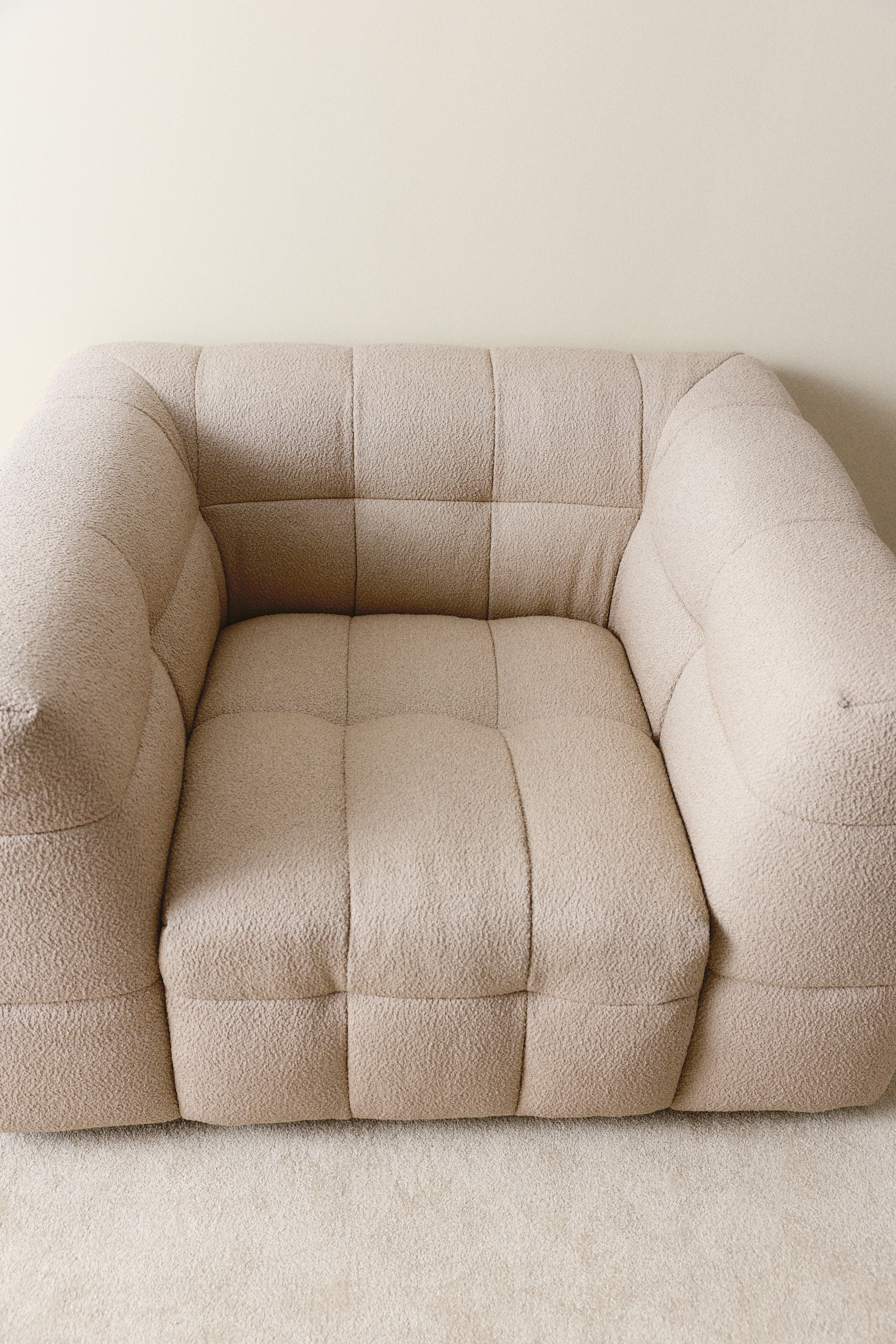 Se større billede: En lys beige bouclé lænestol har et blokagtigt, blødt design med synlig quiltning på tværs af ryglæn, sæde og brede armlæn.