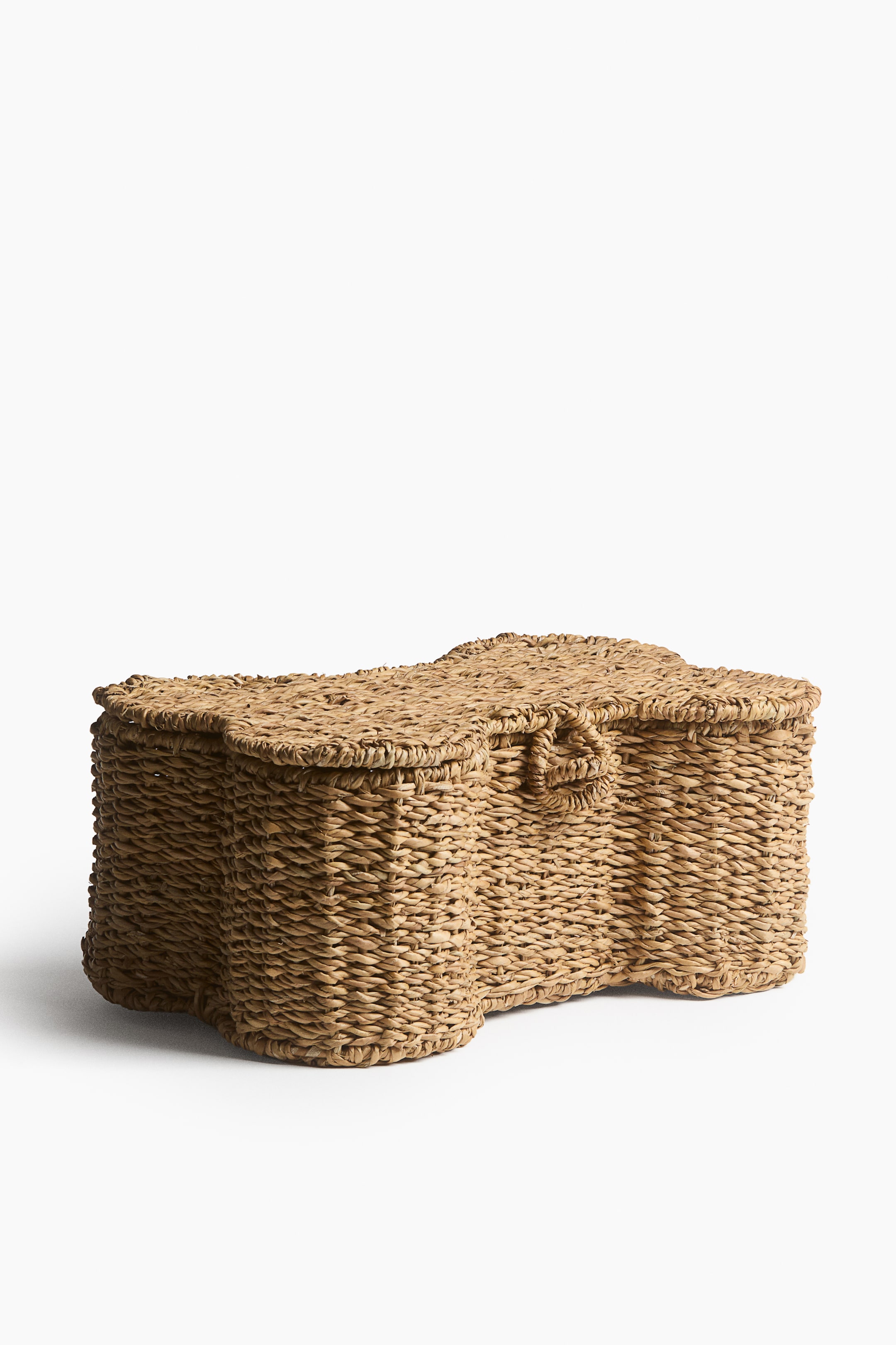 View larger image: A dog-bone-shaped woven storage basket is placed at a slight angle to the front, showcasing its textured natural fibre construction and contoured silhouette. The basket features a matching fitted lid with a small woven loop closure at the centre front.