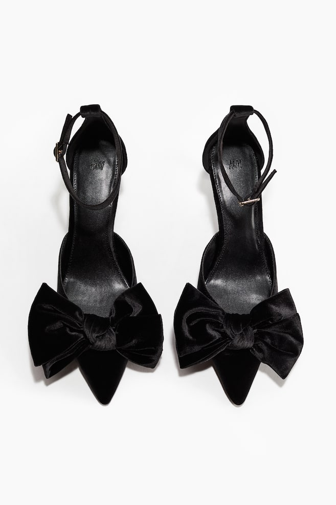 A pair of black velvet D'Orsay style shoes is being presented. Each shoe is featuring a pointed toe, a prominent velvet bow on the vamp, and an ankle strap with a gold-toned buckle. - 1