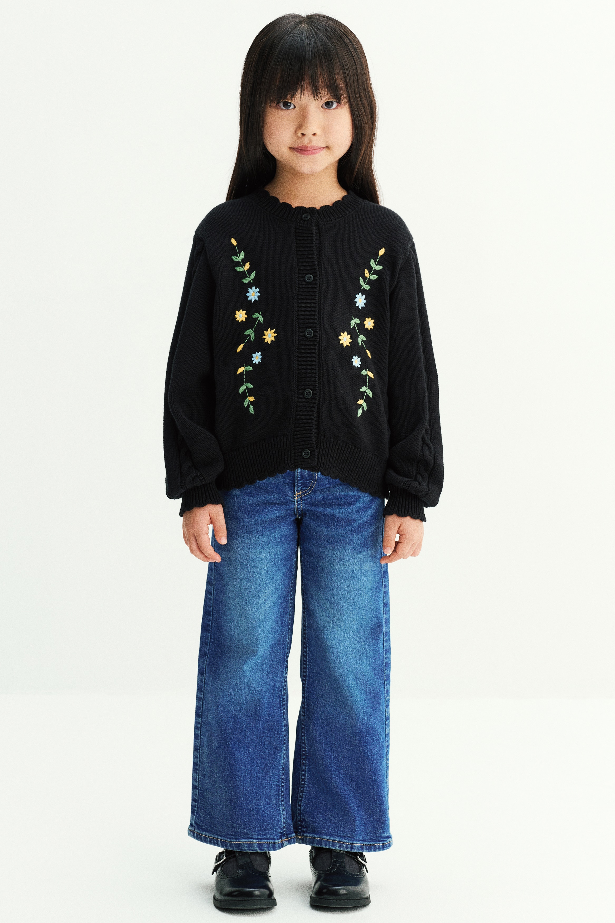A black knit cardigan is shown from the front, featuring colorful embroidered floral vines on either side of the center button closure. The cardigan has a round neckline, dropped shoulders with cable-knit sleeves, and scalloped, ribbed trims along the collar, cuffs, and hem.
