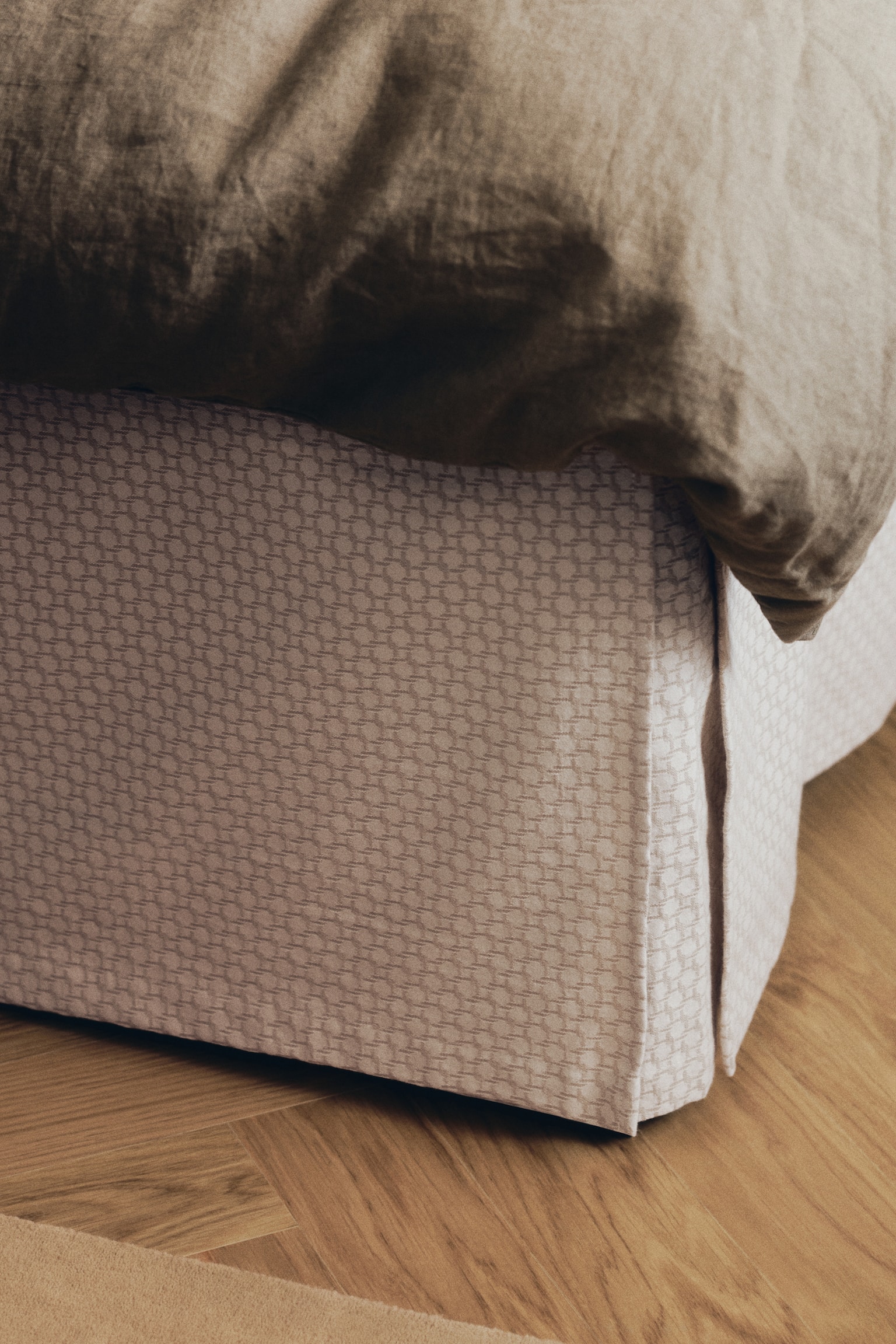 A bedskirt in patterned cotton jacquard is hanging from a bed, showcasing a subtle woven geometric pattern in a light neutral tone. The bedskirt features a structured box pleat at the corner, with a dark olive green duvet resting along the top edge. - 1