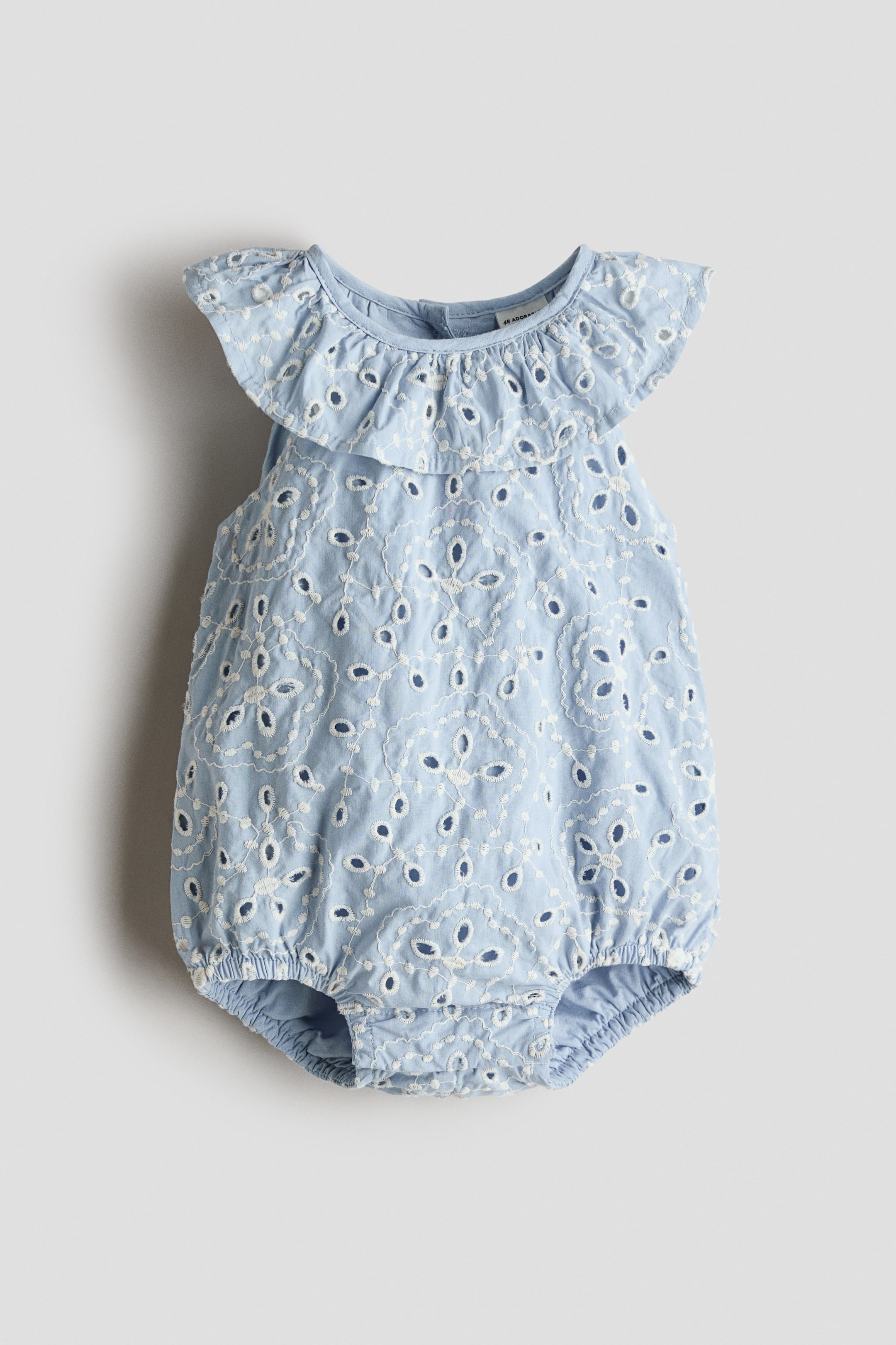 View larger image: A light blue baby romper features white eyelet embroidery patterns across the front, with a ruffled collar extending over the shoulders and elasticated leg openings shaping the bottom.