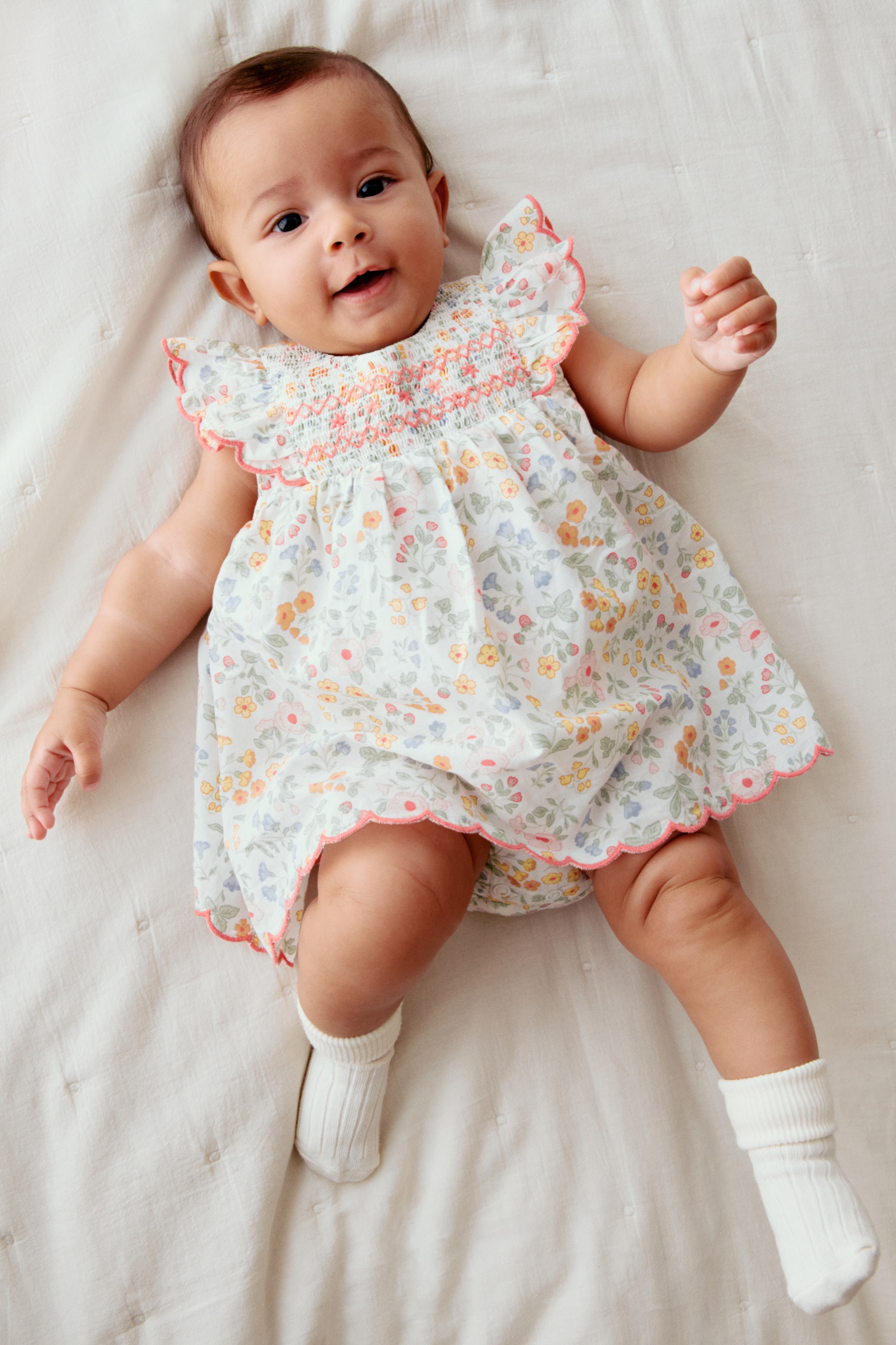 Agrandir l'image: Une robe de bébé avec une base blanche présente un imprimé floral coloré, présentant un corsage smocké avec des coutures couleur pêche-orange et des manches à volants. La jupe et les shorts intégrés de style bloomer sont bordés d'une garniture festonnée assortie couleur pêche-orange.