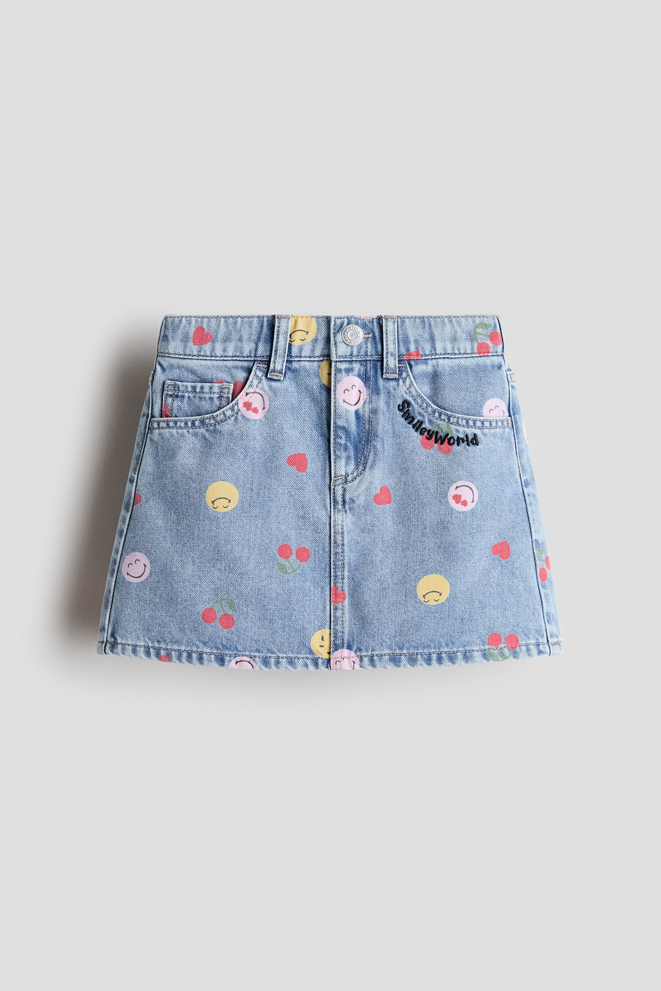 View larger image: A light wash denim skirt is presenting its FRONT view, displaying an A-line shape with a waistband, belt loops, a button and zipper closure, and two front pockets including a small coin pocket. The skirt is patterned with an allover print of various colored smiley faces, red hearts, and cherries, and "SmileyWorld" is embroidered in black on the right front pocket.