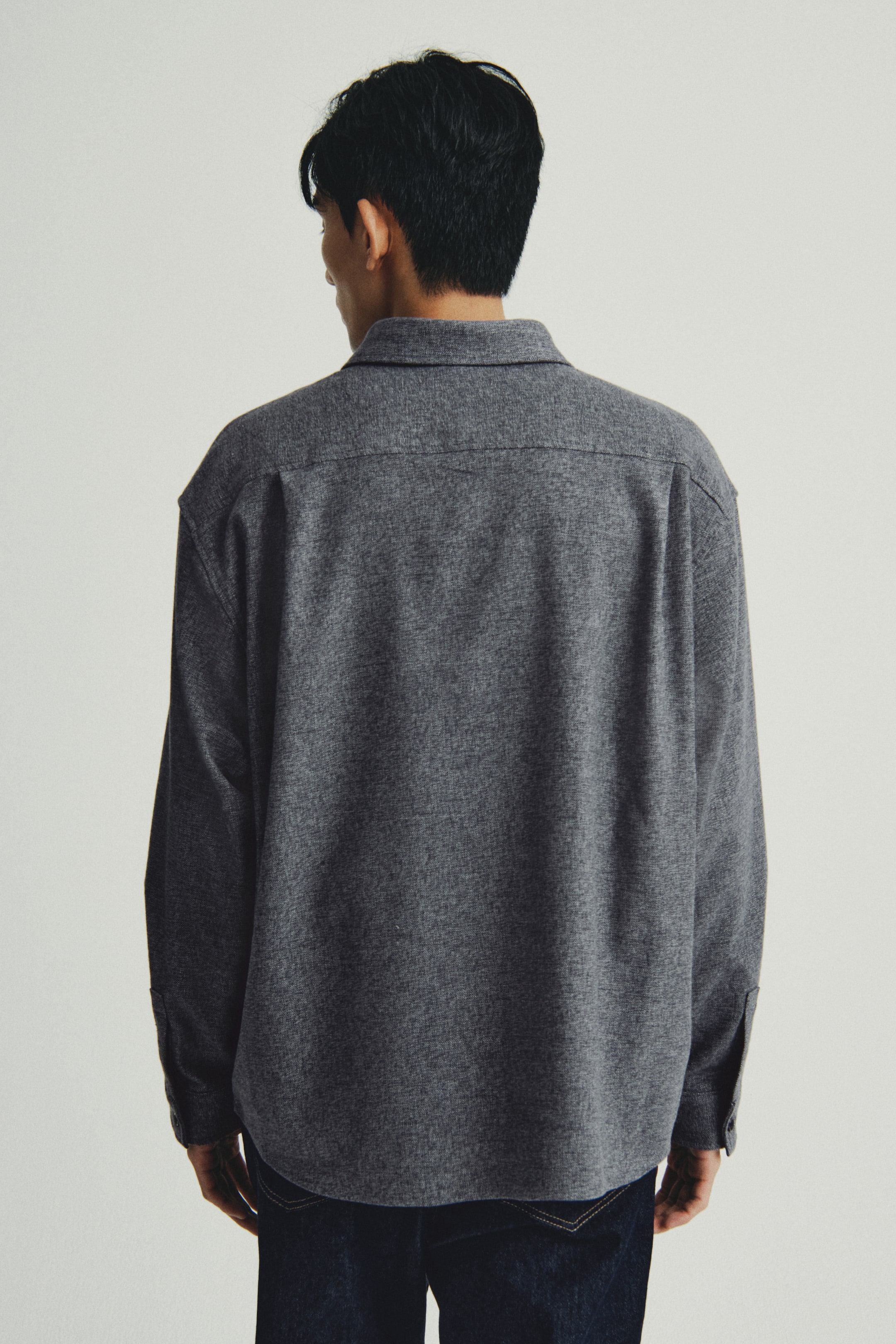 이미지 확대 보기: Heathered 짙은 회색 셔츠가 뒤에서 약간 오른쪽으로 채도되게 보여집니다. 이 셔츠는 포인트 칼라, 옆선 트임, 그리고 버튼된 커프스를 가진 긴 소매가 특징입니다. 이 셔츠는 여유로운 핏과 굽은 밑단을 보여주며, 짙은 색의 청바지를 부분적으로 덮고 있습니다.