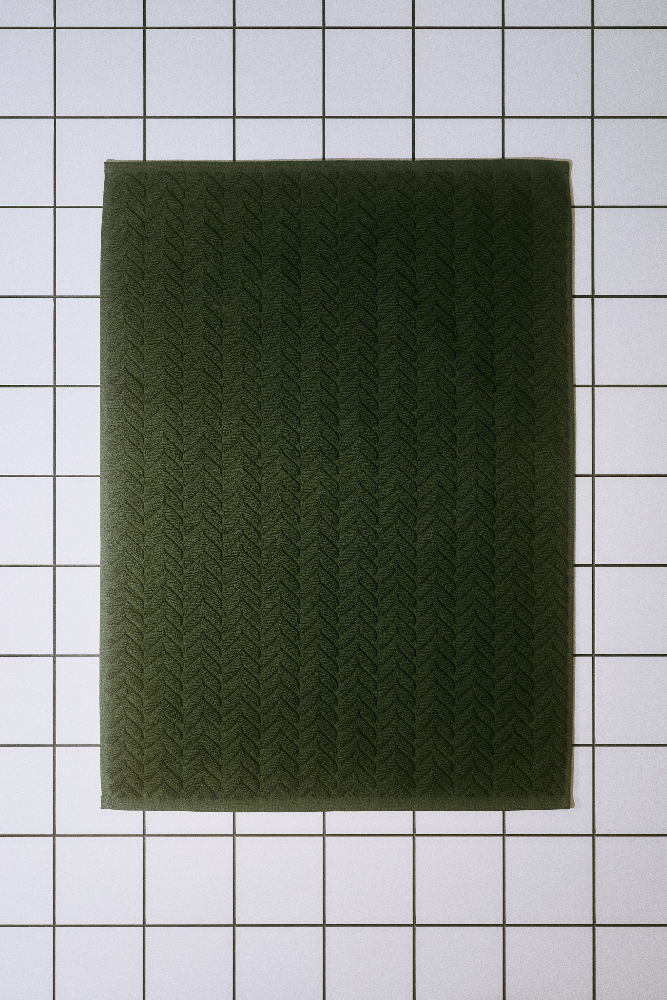 View larger image: A rectangular dark green bath mat features a textured, cable-knit-inspired chevron pattern across its soft cotton terry surface. The mat has a flat, solid border along its outer edges.