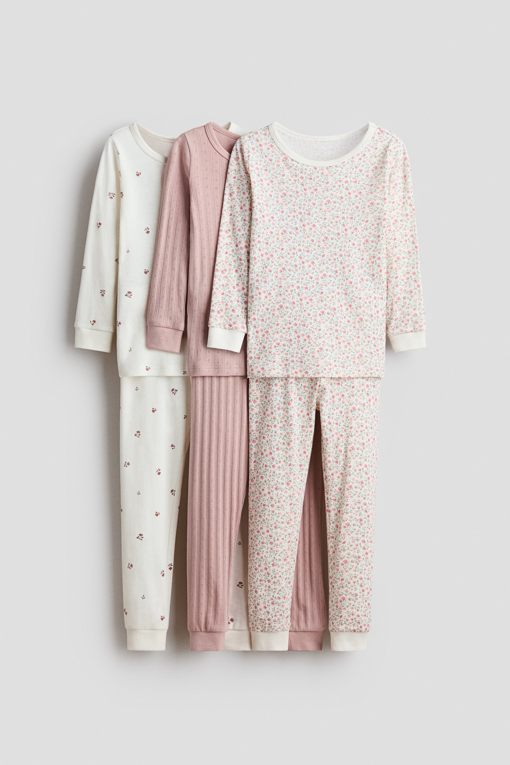 View larger image: Three pajama sets are displayed side-by-side, each featuring a long-sleeve top and matching long pants with rib-knit cuffs. The sets include an off-white one with scattered reddish-brown motifs, a solid dusty rose pink one with a fine rib-knit texture, and an off-white one with a dense pink floral and green leaf pattern.