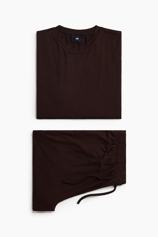A set of dark brown pyjamas is presented, featuring a neatly folded T-shirt and a pair of folded shorts. The T-shirt lies flat, showing its front with a round, rib-trimmed neckline, while the shorts display an elasticated, drawstring waistband with a visible drawstring tie. - 1