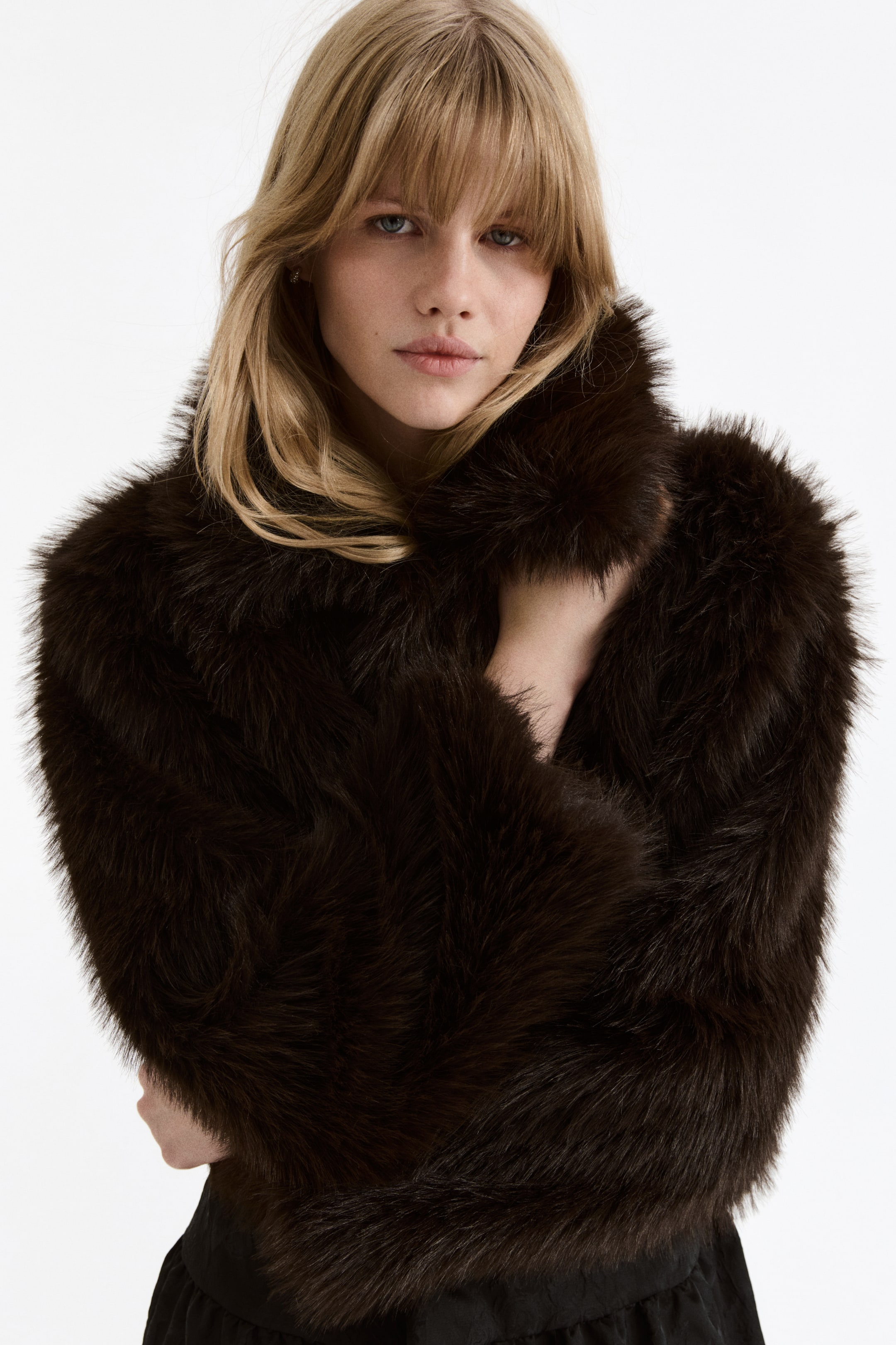 View larger image: A dark brown faux fur jacket is featuring a high, plush collar that frames the face. Its shaggy texture covers the shoulders and arms, with one hand holding the front to highlight its enveloping design.