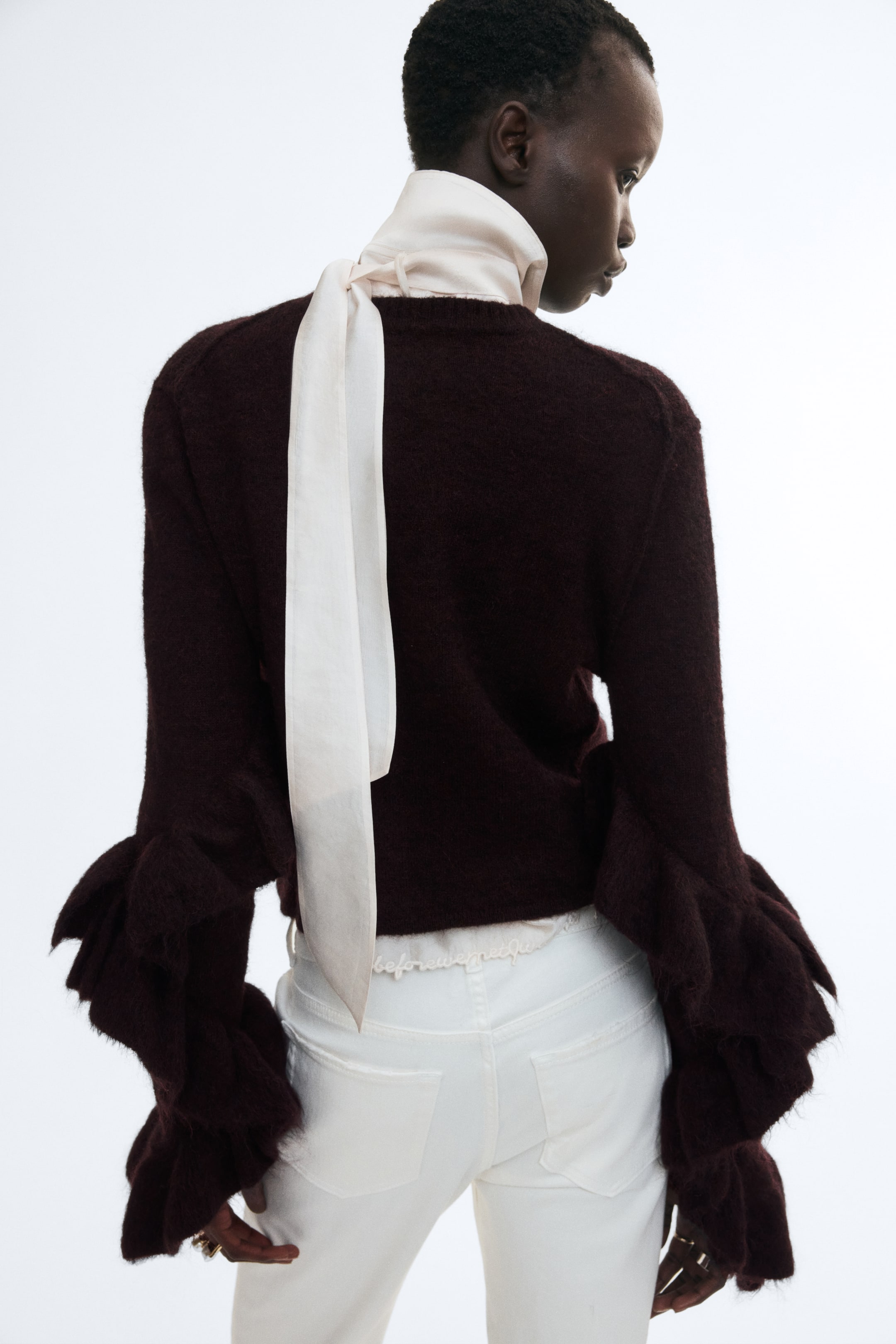 Agrandir l'image: Un cardigan en maille bordeaux foncé en mélange de mohair et de laine est porté par le mannequin, présenté de dos. Le cardigan présente d’imposants volants à étages sur le bas des manches et une base côtelée, et il est associé à une écharpe drapée de couleur crème ainsi qu’à un pantalon blanc.