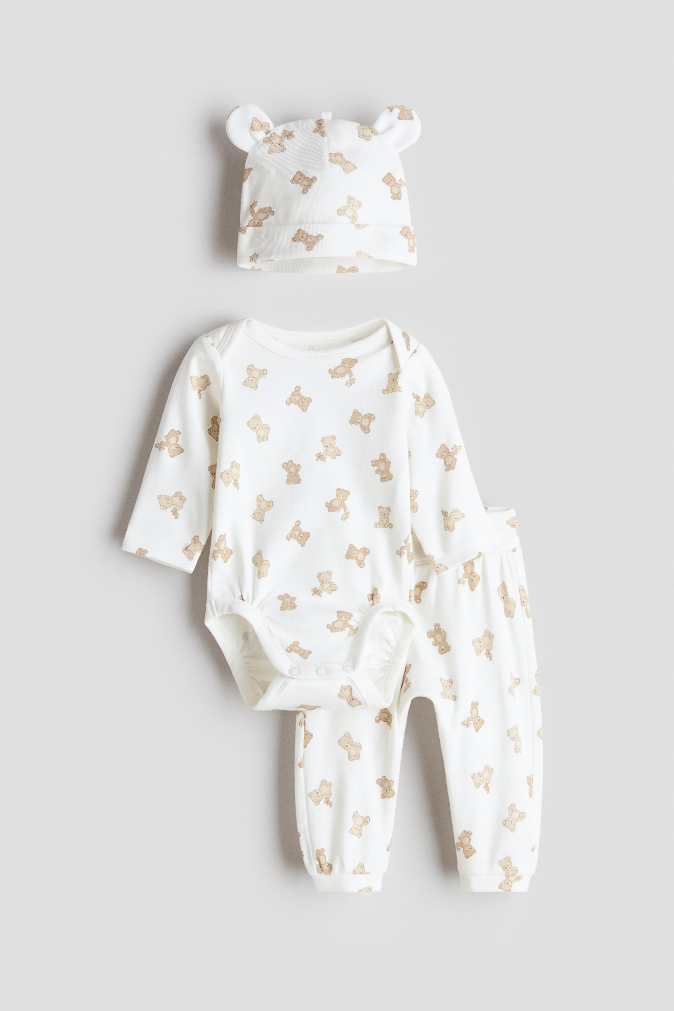 View larger image: A three-piece baby clothing set features a white bodysuit, trousers and a hat, all patterned with brown teddy bears. The front-facing bodysuit displays a round neckline, long sleeves, press-studs at the bottom and gathered leg seams. The accompanying trousers have a foldover waist and cuffed hems, while the hat features small ear details.