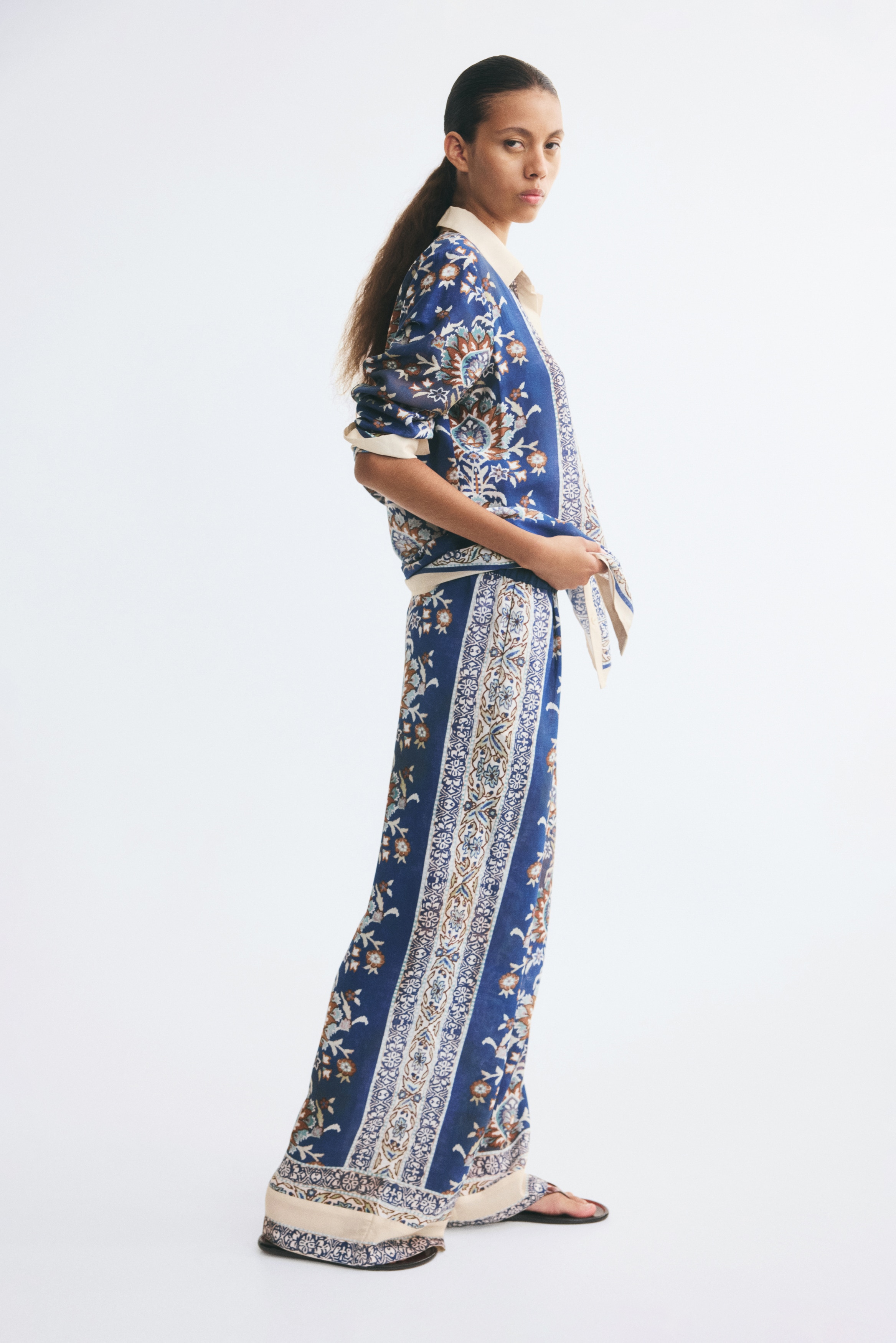 These wide-leg, loose-fit trousers are shown from the front, featuring a high, elasticated waistband and an ornate floral and border pattern in blue, white, and brown. The trousers are displayed with wide legs and a contrasting cream-coloured patterned band at the hems.