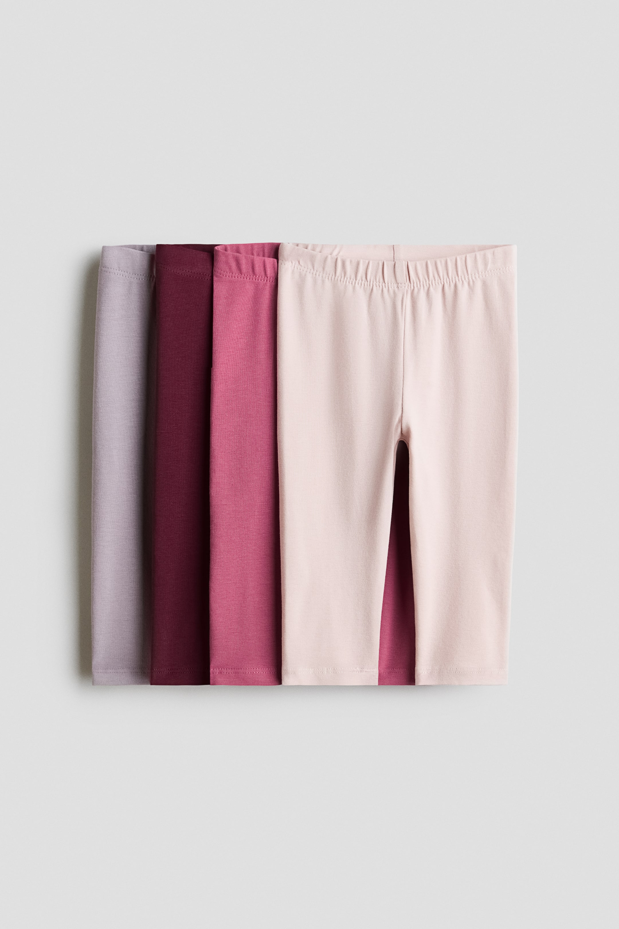 View larger image: Four pairs of cotton jersey leggings are layered together, with the front pair in light pink facing forward to show its covered elasticated waistband and straight-leg design. The other three pairs are stacked behind, displaying shades of dusty rose, dark burgundy, and light purple.