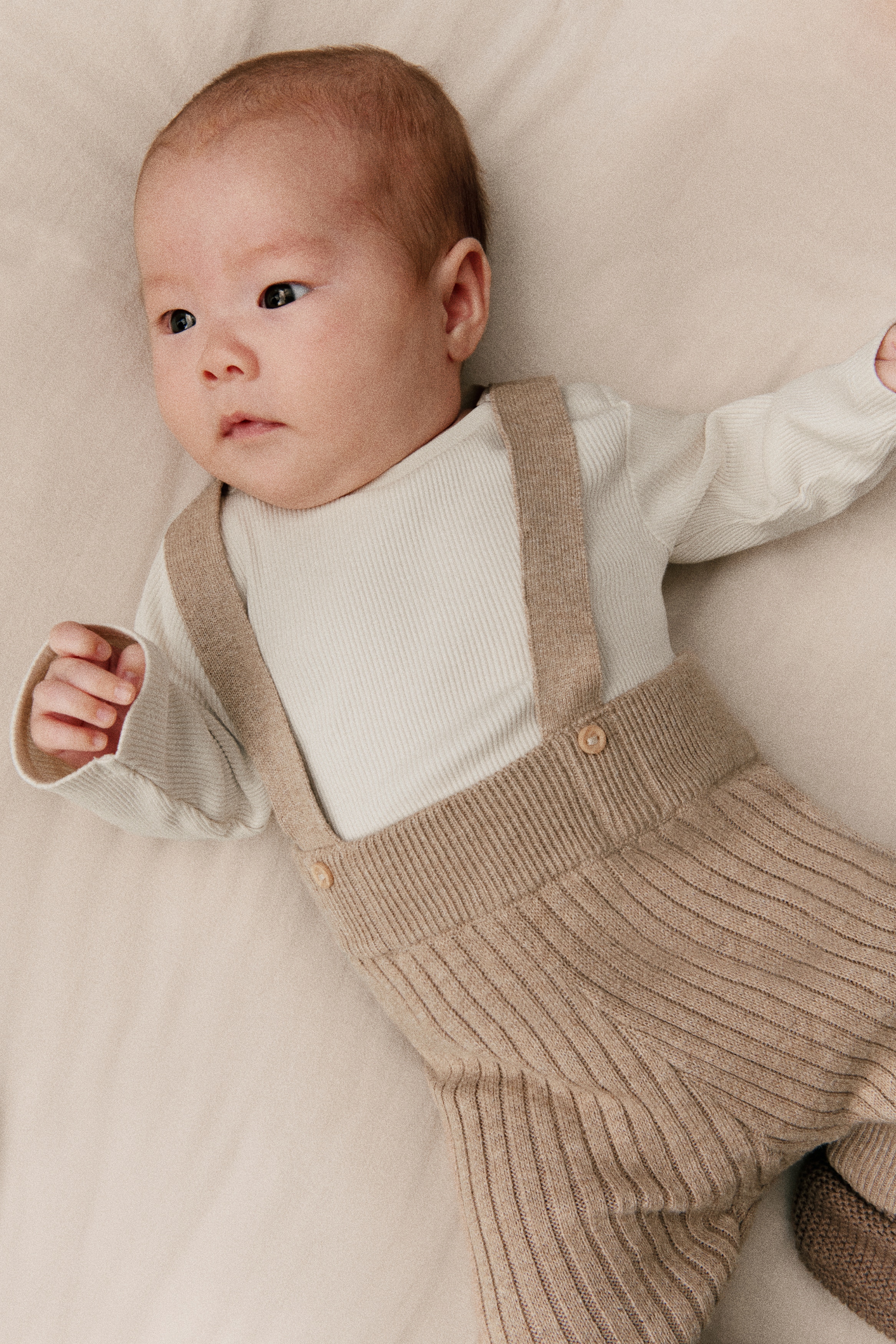 A baby outfit is being presented from the front, featuring a white long-sleeved top with a subtle ribbed texture. Beige knitted dungarees are being worn over the top, showcasing a prominent vertical rib knit pattern. The dungarees' shoulder straps are fastening at the front with two light wooden buttons, and the leg cuffs are folded up with a ribbed hem.