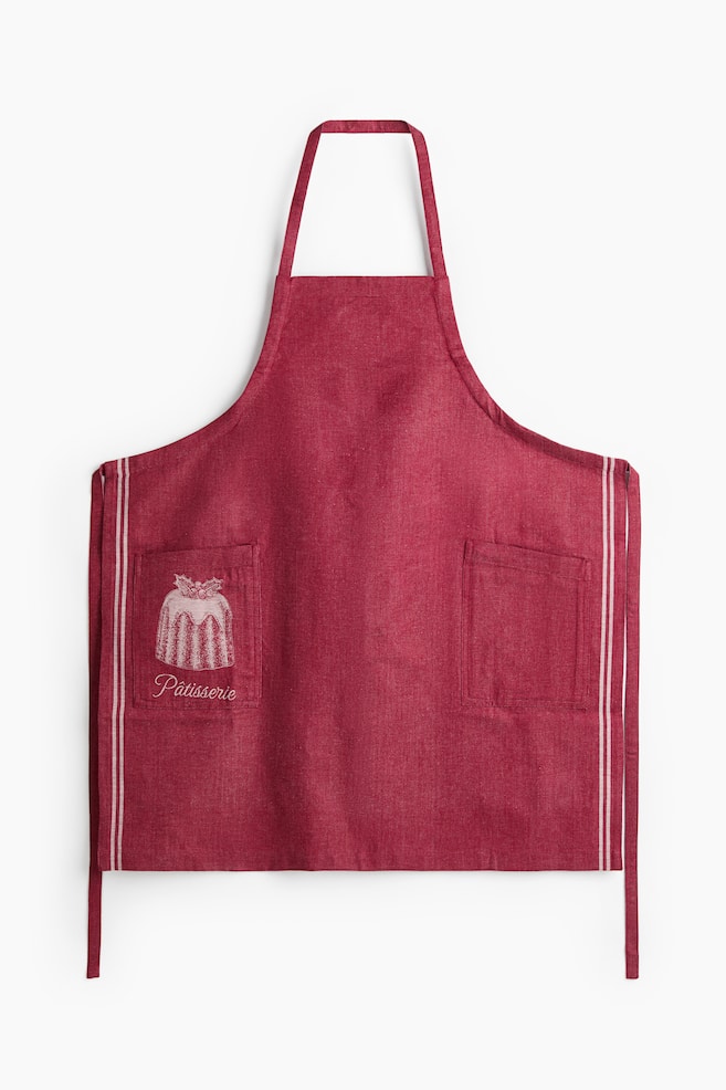 A front-facing red bib apron featuring textured fabric, a neck loop and side waist ties. It has two front patch pockets with thin vertical white stripes running down the outer edges, while the right pocket displays a white graphic of a bundt cake with the word "PÃ¢tisserie" printed underneath. - 1
