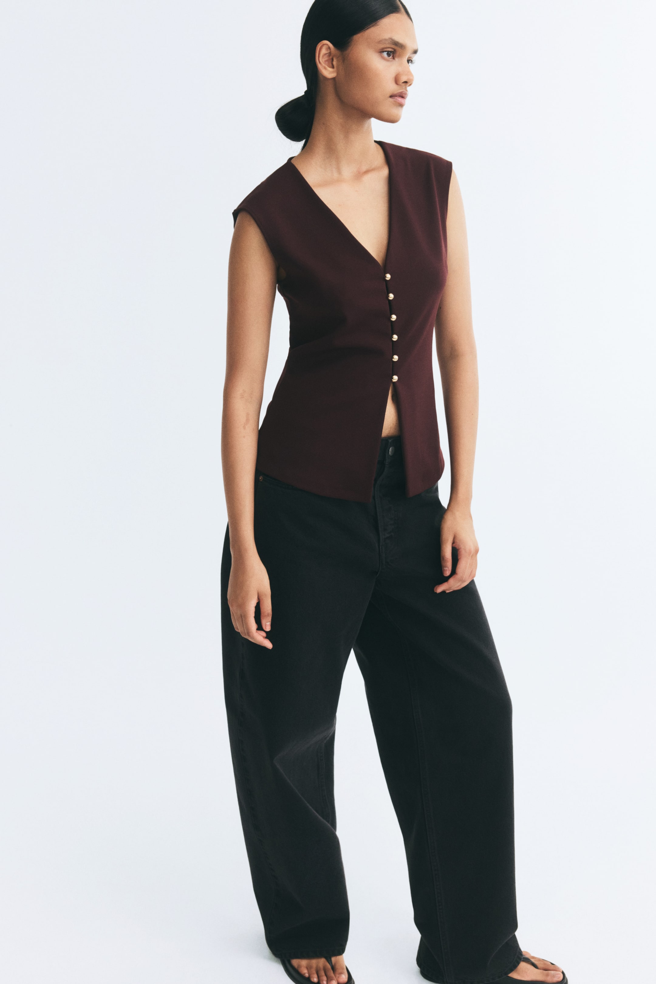 View larger image: A dark burgundy vest in ponte fabric is being worn by the model, featuring a deep V-neckline and a row of gold-toned, domed metal buttons down the front. The sleeveless vest is styled with wide-leg black trousers.