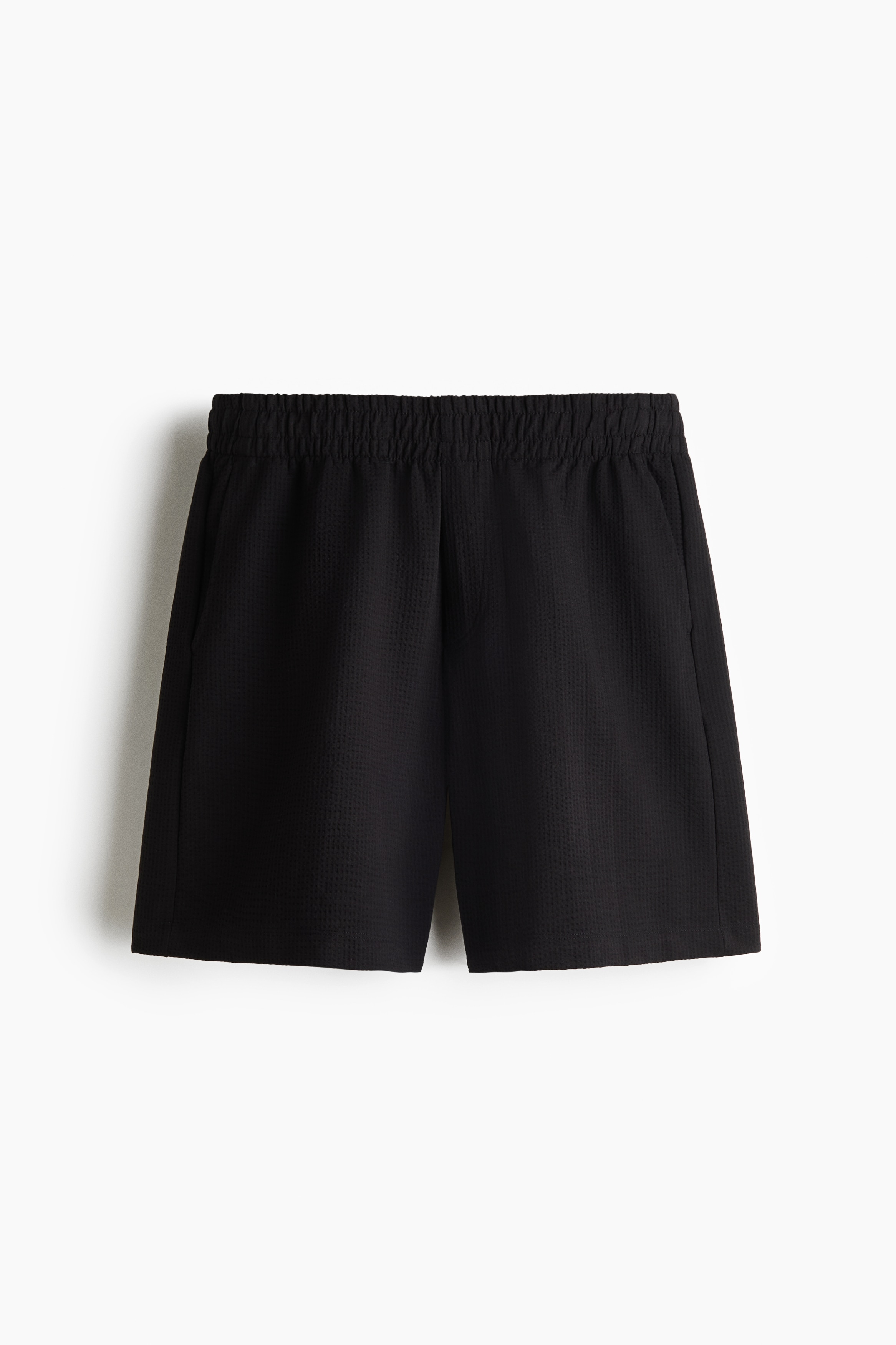 Chiáº¿c quáº§n short dÃ¡ng lá»­ng báº±ng cháº¥t jersey gÃ¢n Ã´ vuÃ´ng mÃ u tá»i, cÃ³ phom rá»ng thoáº£i mÃ¡i vÃ  cáº¡p thun co giÃ£n, Äang ÄÆ°á»£c ngÆ°á»i máº«u máº·c, vá»i má»t tay Äáº·t trong tÃºi bÃªn hÃ´ng. Quáº§n short ÄÆ°á»£c phá»i cÃ¹ng Ã¡o sÆ¡ mi tay ngáº¯n Äá»ng bá», máº·c má» bÃªn ngoÃ i Ã¡o tank top mÃ u xÃ¡m ÄÆ°á»£c sÆ¡ vin gá»n gÃ ng.
