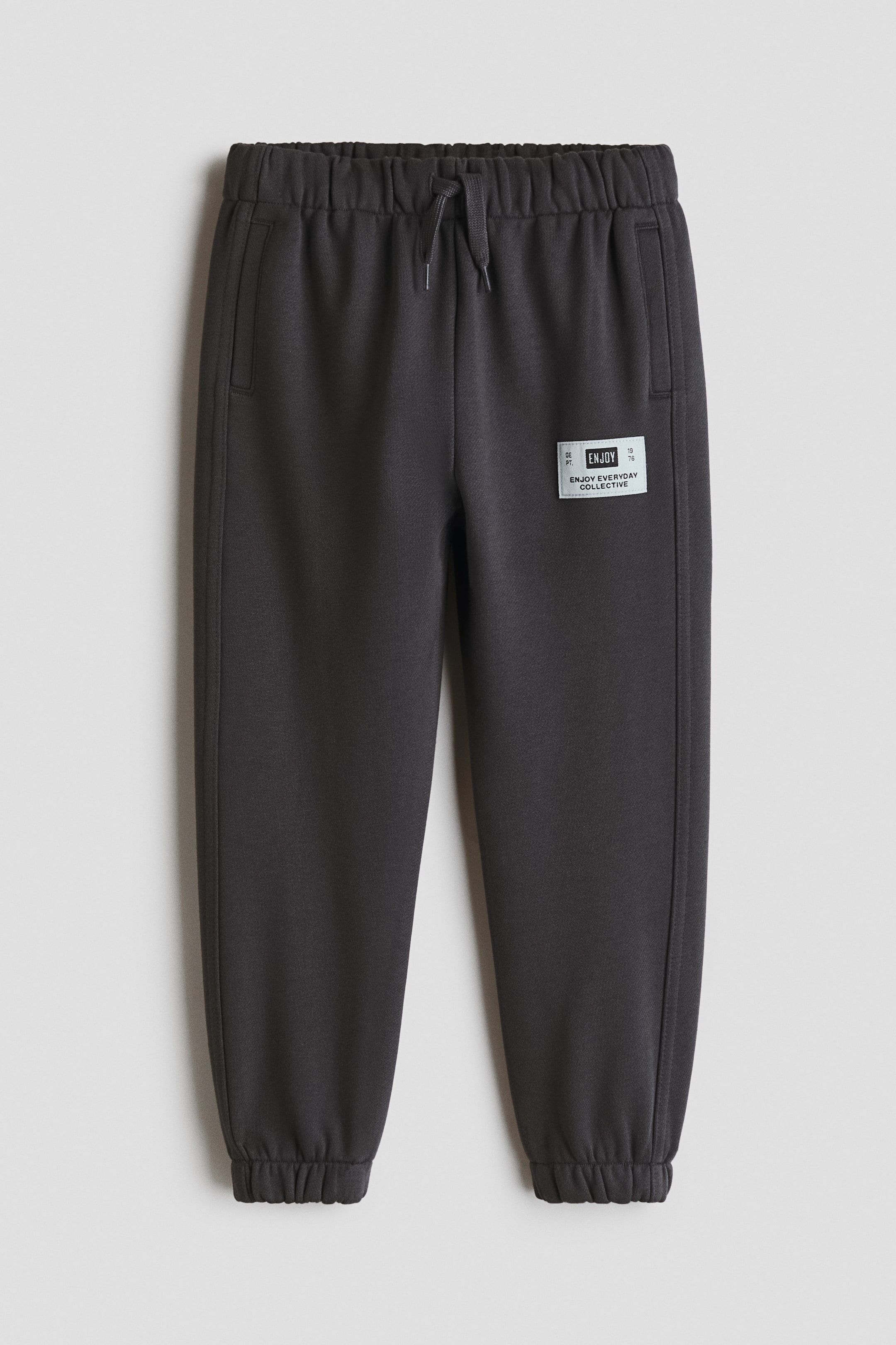 Ampliar la imagen: Joggers gris oscuro, hechos de tela ligera de sudadera, se presentan desde el frente. Cuentan con una cintura elástica con cordón, bolsillos laterales, dobladillos elásticos y un parche rectangular en la parte superior de la pierna derecha.