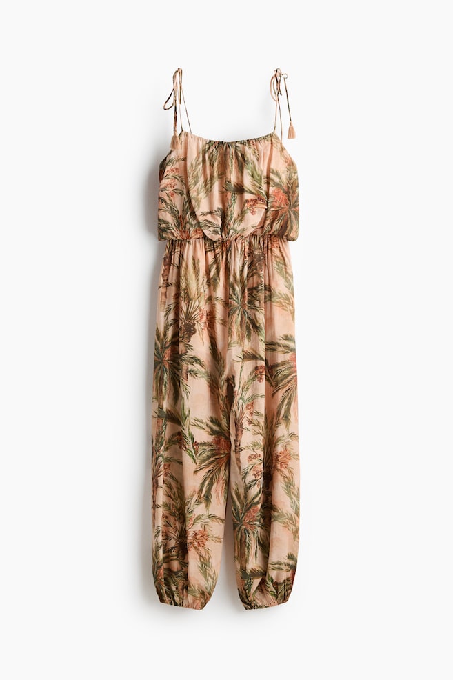 A front-facing jumpsuit featuring a light peach base adorned with a tropical palm tree print. It is designed with thin spaghetti straps tied at the shoulders with tassels, an elasticated blouson bodice, an elasticated waist, and loose-fitting legs gathered at elasticated cuffs. - 1