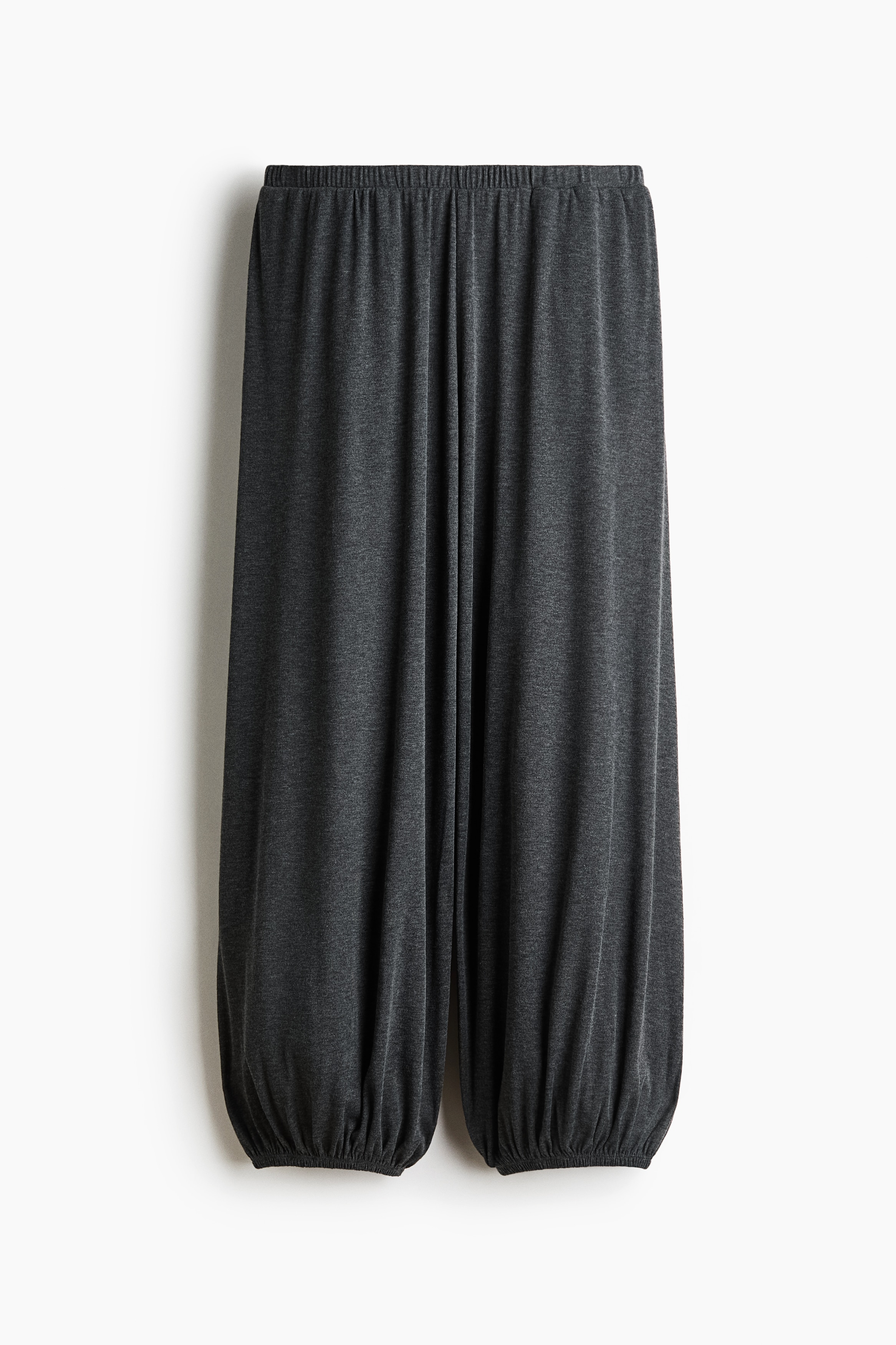 Relaxed-fit jersey trousers in dark grey, featuring balloon legs with elasticated hems, are worn by the model with her hands in the side pockets. The trousers are styled with a matching short-sleeve top tied at the waist and black sandals.