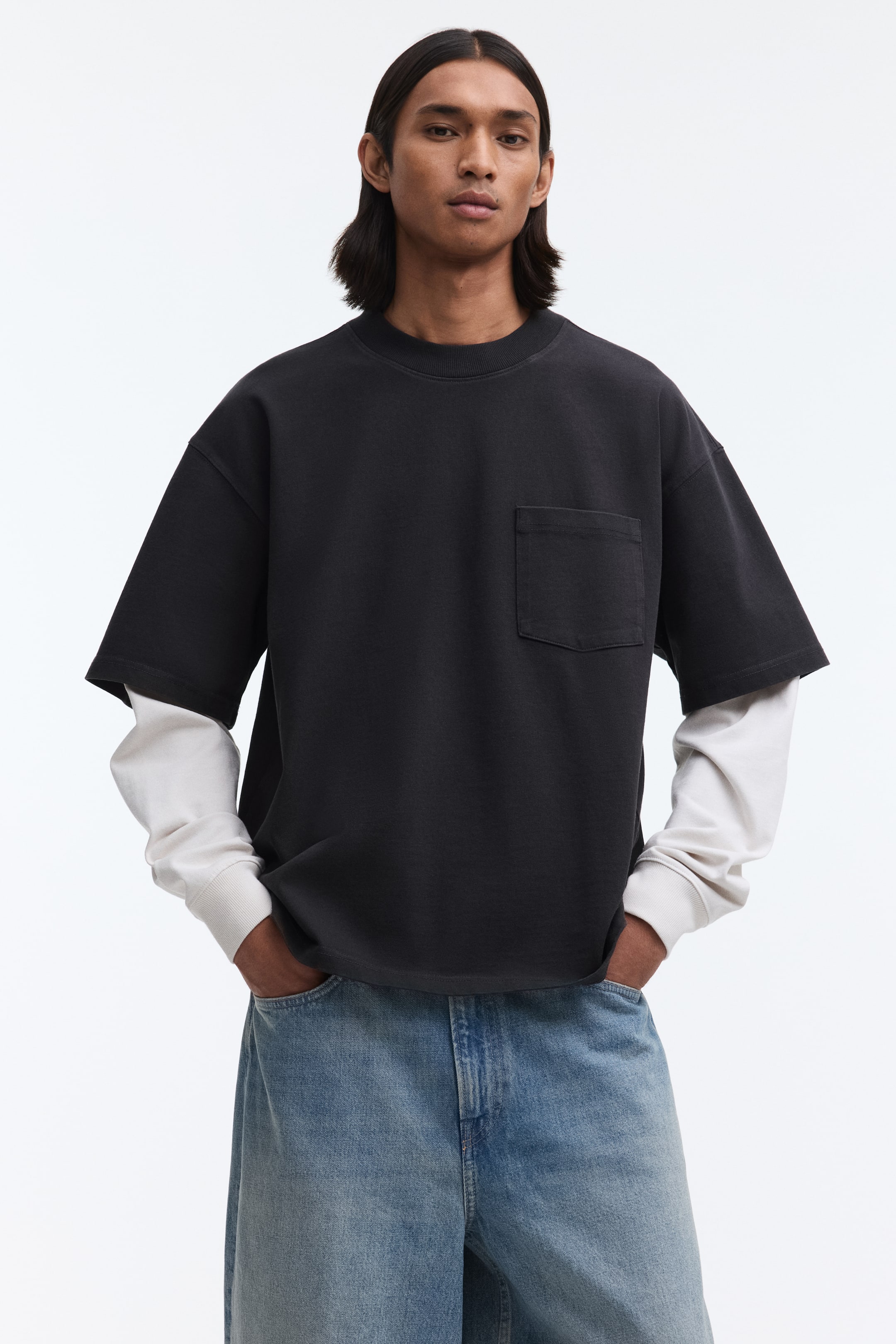 View larger image: The dark gray oversized T-shirt features a crew neck and a patch pocket on the left chest. It showcases dropped shoulders and short sleeves, worn layered over a light-colored long-sleeved shirt.